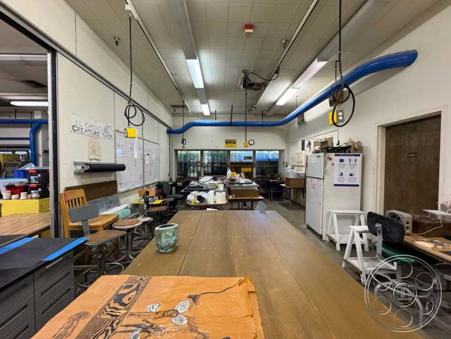 Old School - task chairs, large windows providing natural light, bright fluorescent ceiling lights, metal storage cabinets, interior workshop space, exposed steel ceiling beams, industrial-grade concrete flooring, blue industrial ventilation pipes, wooden worktables with natural finish, open layout with multiple workstations, utility room with refrigerator, cluttered creative workspace, dry-erase boards on walls, overhead electrical cables, paint splattered surfaces, hanging tools and equipment, artistic mural on wall