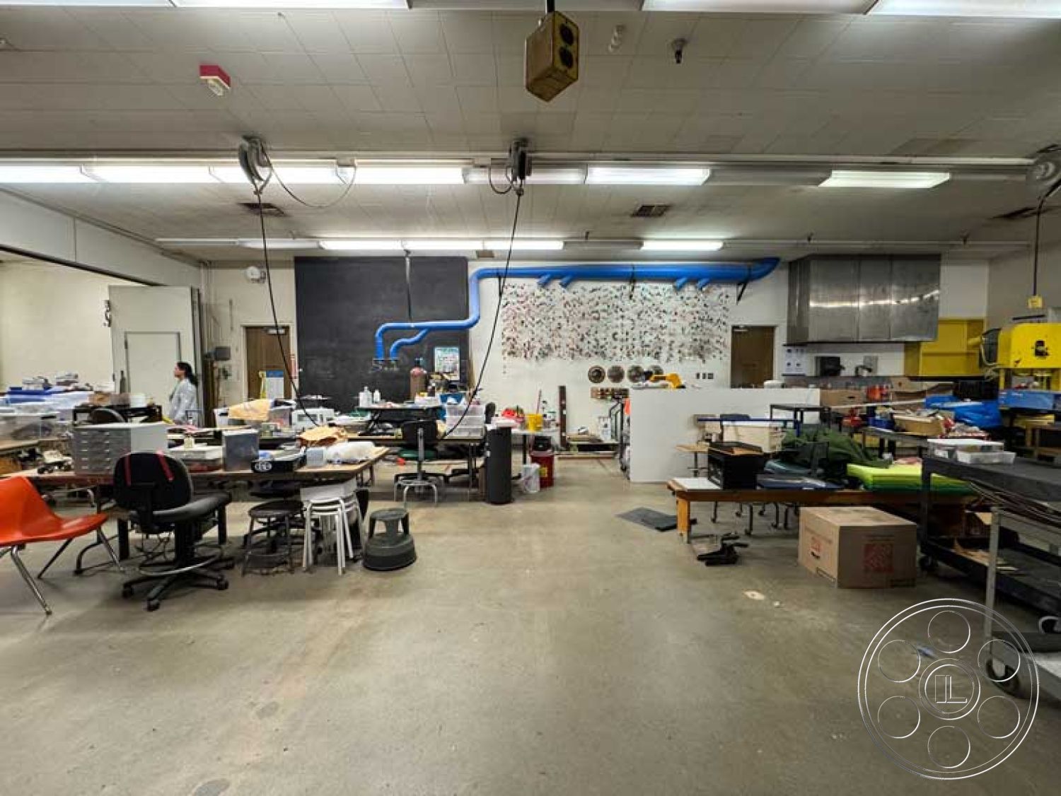 Old School - artistic wall decor, metal shelving units, suspended lighting fixtures, exposed concrete floor, creative studio space, white painted walls, urban industrial setting, black chalkboard wall, utility sink area, industrial workshop interior, rolling tool cart, blue PVC pipe installation, various work tables, task chairs in bright colors, organized cluttered workspace, overhead storage, multifunctional workspace design, open-layout workshop, functional workshop equipment, contemporary workspace aesthetic