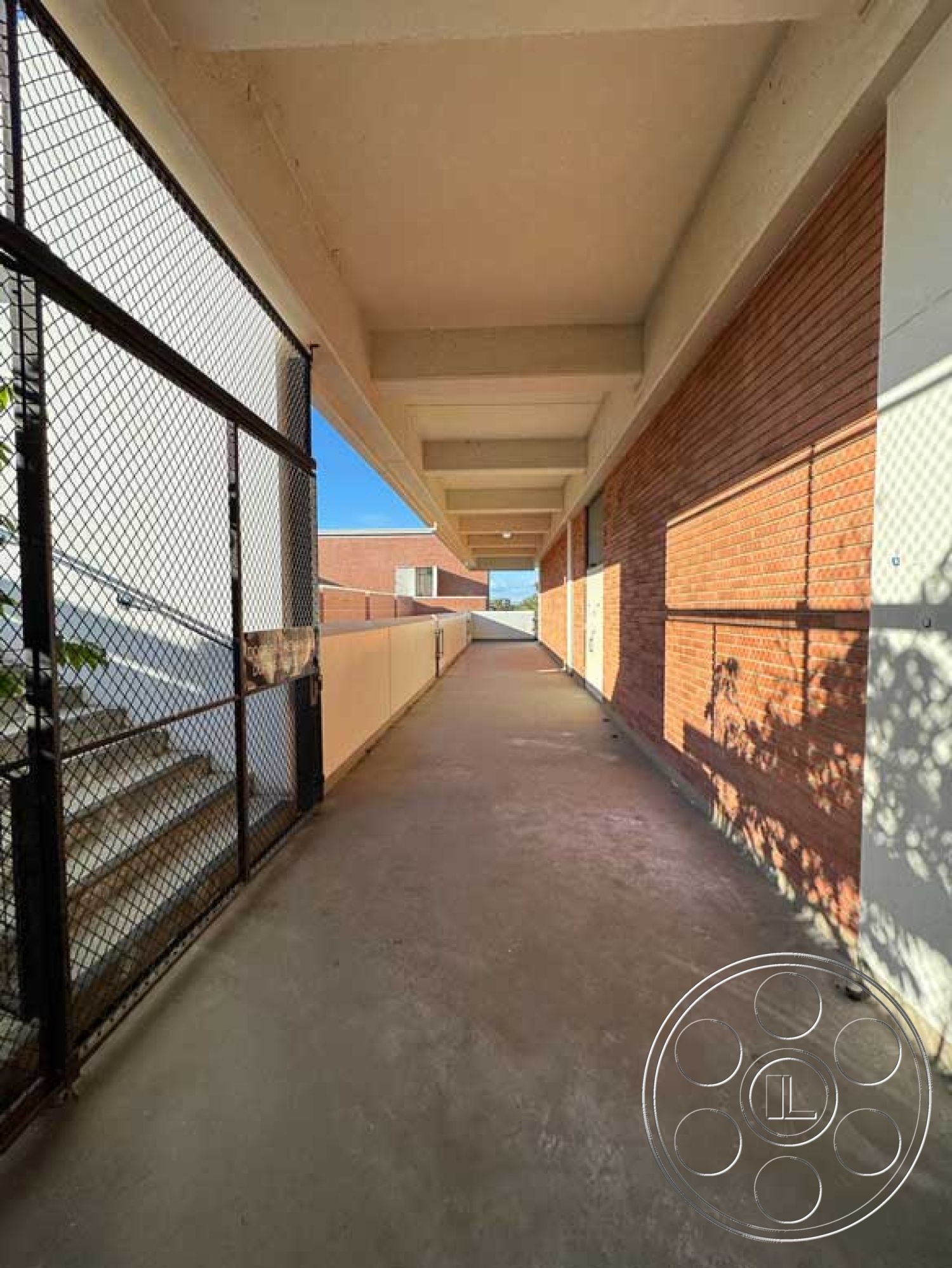 Old School - industrial aesthetic, textured concrete surface, modern architectural design, minimalist architectural details, straight corridor layout, interior corridor space, brick building facade, brick exterior walls, metal chain-link fencing, exposed concrete ceiling beams, urban school environment, composite concrete walkway flooring, natural lighting from overhead windows, structural concrete elements, outdoor covered walkway