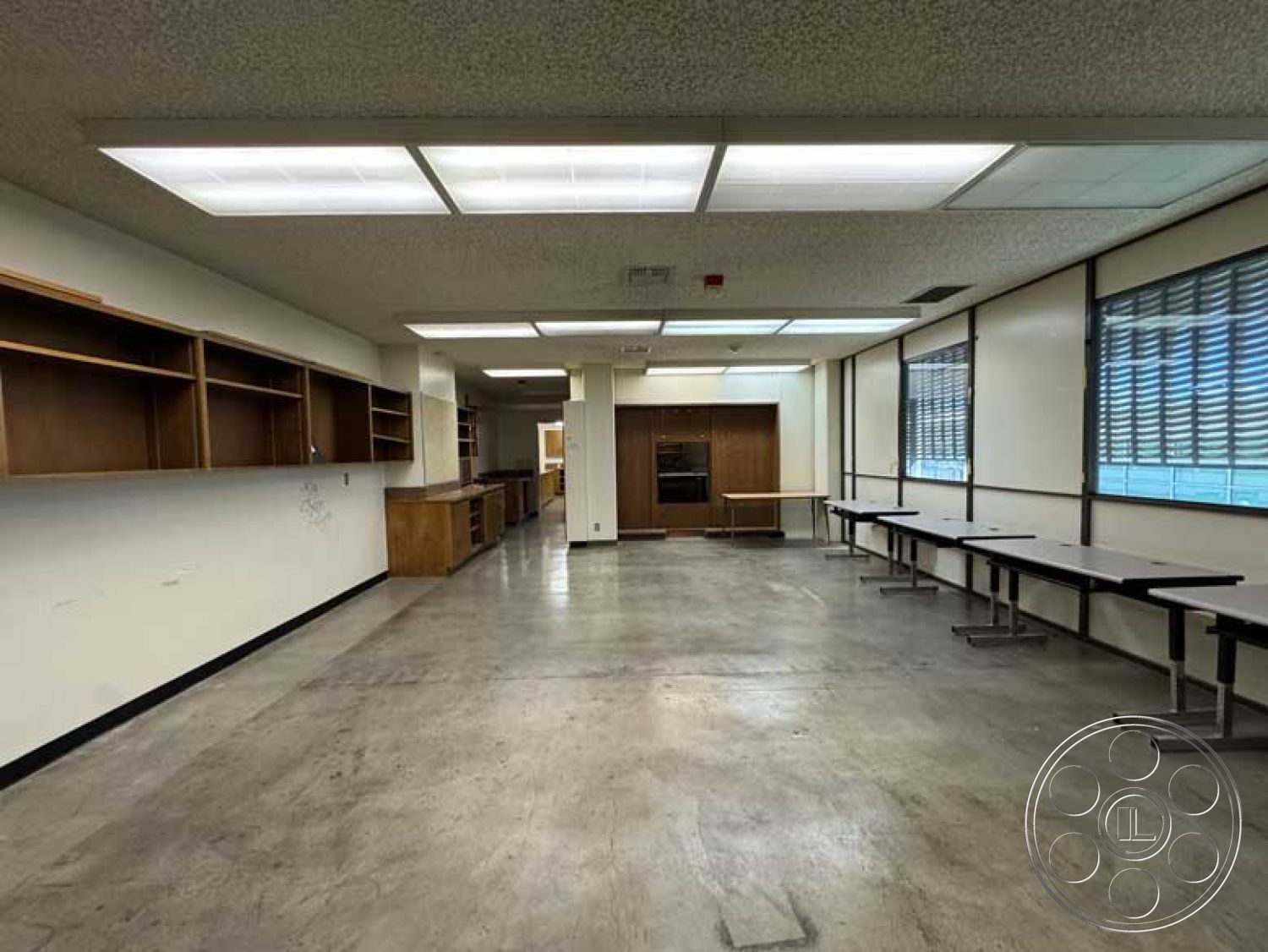 Old School - neutral wall color, minimalist design aesthetic, open floor plan layout, fluorescent ceiling lights, interior concrete floor, large windows with blinds, exposed wooden cabinets, linear wall-mounted shelves, office space with modular tables