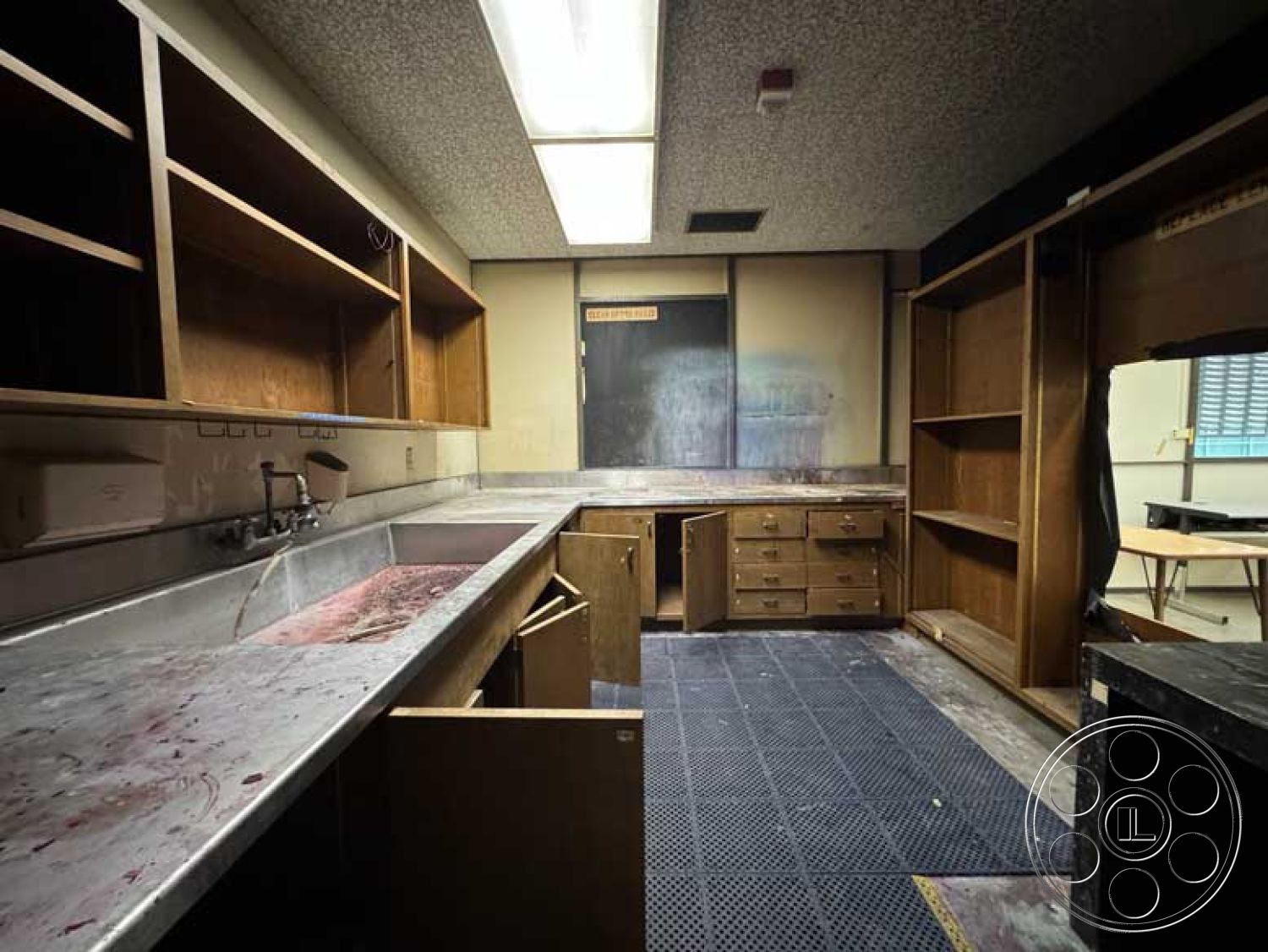 Old School - fluorescent ceiling lights, wood cabinetry with open shelving, interior kitchen with stainless steel countertops, tile flooring with rubber flooring sections, dark wooden storage cabinets, double sink kitchen area, modern industrial style design, kitchen layout with L-shaped counter, dirty and worn surfaces, basic functional kitchen space, utilitarian workspace environment