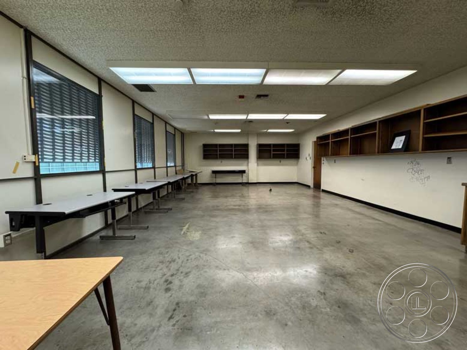 Old School - concrete flooring, large windows for natural light, task lighting, industrial design elements, acoustic ceiling tiles, drop ceiling with fluorescent lights, multifunctional room, interior classroom space, modern educational environment, light gray wall paint, built-in wood shelving, steel frame windows, square classroom layout, exposed electrical fixtures, open space configuration