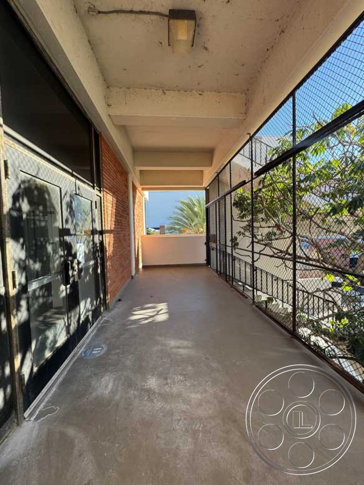 Old School - modern architectural details, natural light from doorway, exposed concrete ceiling beams, brick exterior wall, concrete walkway floor, black metal security door, fenced balcony with metal grid, urban corridor with landscaping views, interior access to outdoor space, city setting with palm trees