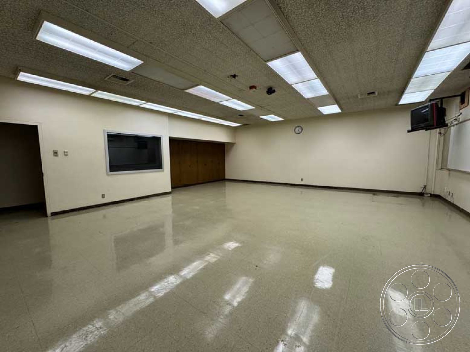 Old School - vinyl flooring, plain white walls, wood paneling accent wall, drop ceiling with fluorescent lights, clock on wall, functional educational space, large window frame, empty classroom layout, spacious room design