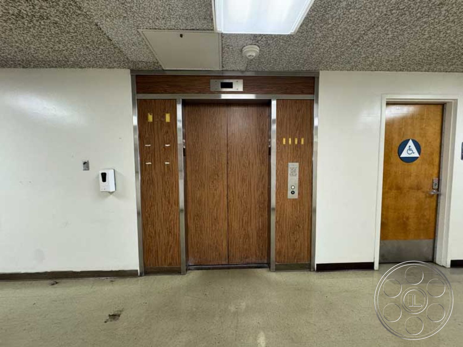 Old School - acoustic ceiling tiles, textured plaster wall finish, white painted walls, minimalist architectural details, modern elevator design, public building interior, interior elevator doors, wood panel elevator facade, stainless steel elevator trim, light fixture ceiling panel, commercial-grade vinyl flooring, handicapped accessible bathroom door, signage for restroom facilities, adjacent hallway space, neutral-toned wall color