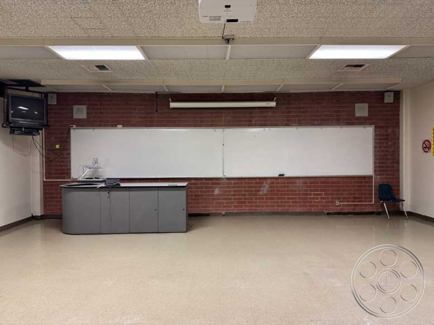 Old School - wall-mounted television, acoustic ceiling tiles, vinyl tile flooring, interior classroom space, light-colored painted walls, exposed electrical outlets, brick classroom walls, whiteboard teaching surface, grey laminate teacher's desk, overhead projector equipment, single blue student chair, classroom lighting fixtures