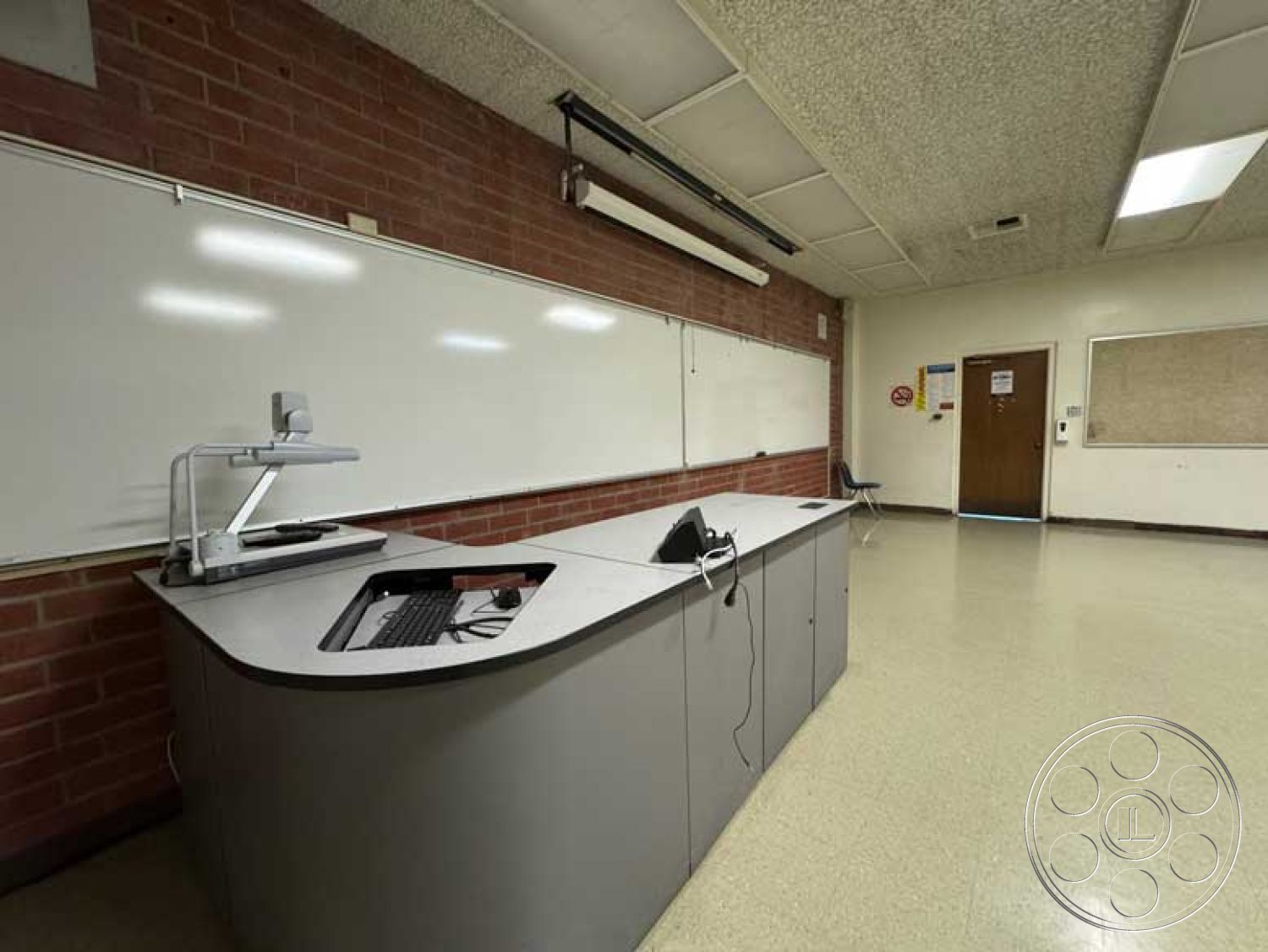 Old School - exposed brick accent wall, drop ceiling with acoustic tiles, fluorescent ceiling lights, interior classroom setting, simple chair design, whiteboard wall treatment, laminate classroom flooring, curved teacher's desk, standard classroom door, built-in cabinets for storage, minimalistic room layout