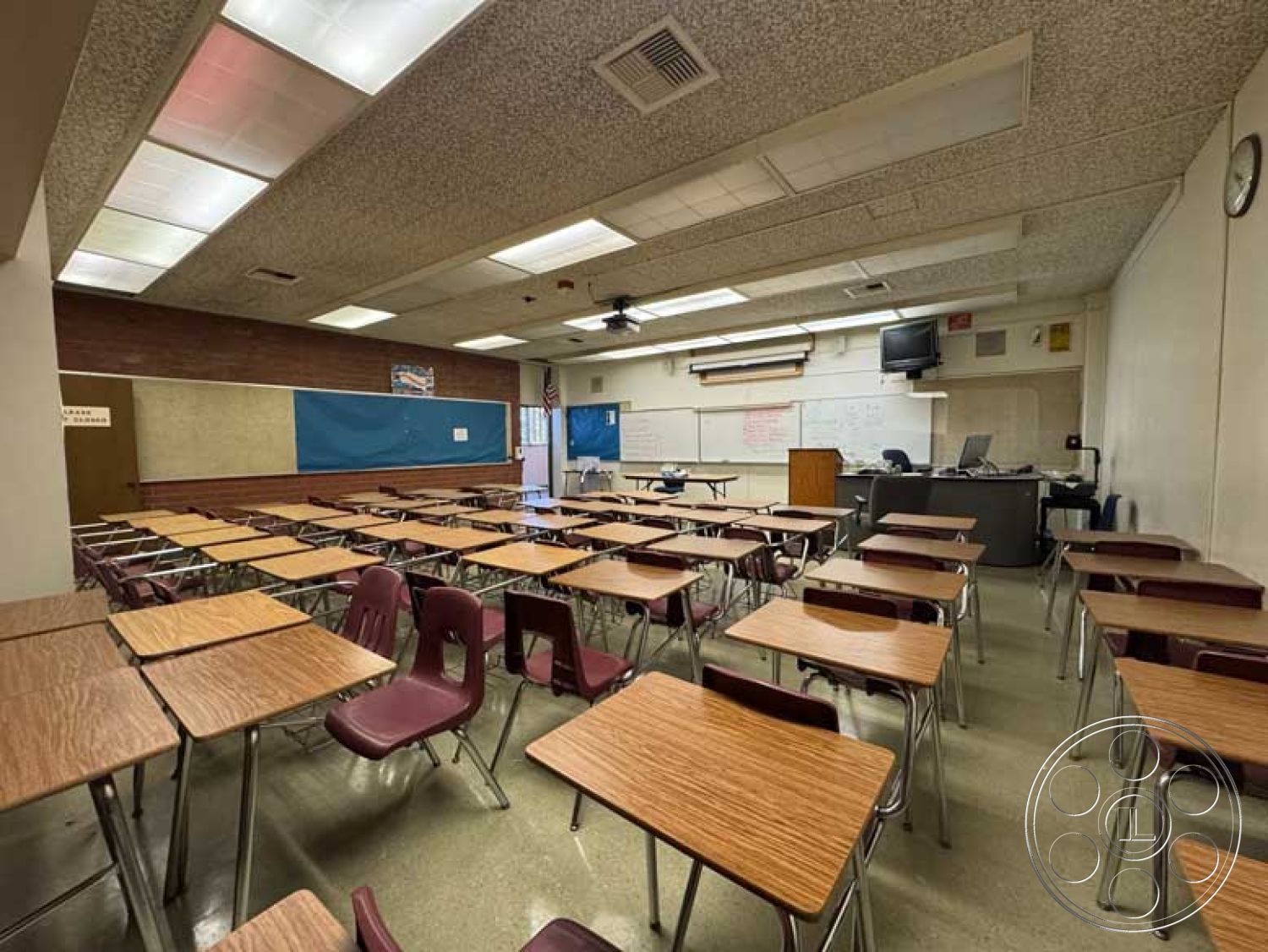 Old School - brick accent wall, overhead fluorescent lighting, textured acoustic ceiling tiles, interior classroom design, dry erase board wall treatment, plastic classroom chairs, large window with blinds, classroom layout with rows of desks, hardwood flooring in classroom, metal student desks, teacher's podium, educational technology setup, chalkboard with blue background, fixed wall mounted projector, built-in classroom storage units, modern educational setting, secondary education environment