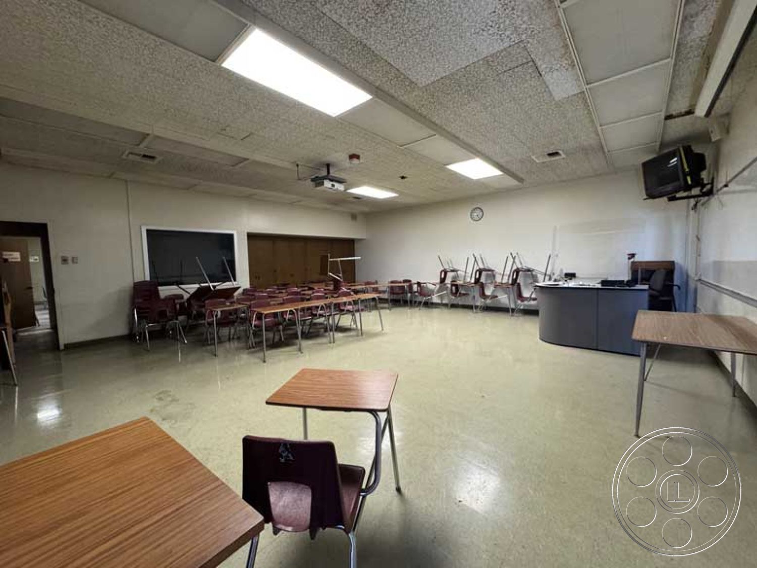 Old School - large windows, wall-mounted television, fluorescent lighting, wooden doors, wall clock, acoustic ceiling tiles, textured plaster wall finish, suspended ceiling tiles, vinyl tile flooring, interior classroom space, wooden teacher's desk, stackable plastic student chairs, projector ceiling mount, room with minimal furniture arrangement, empty learning environment, chalkboard area
