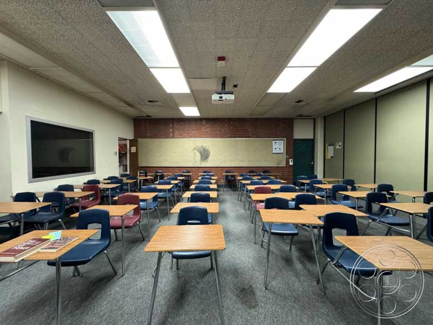 Old School - open floor plan, brick accent wall, vinyl flooring, textured plaster wall treatment, drop ceiling with recessed lighting, interior classroom space, projector mounted ceiling, blue plastic chairs, urban educational setting, metal framed desks, window with black frame, whiteboard on wall, large bulletin board, custom cabinetry with storage