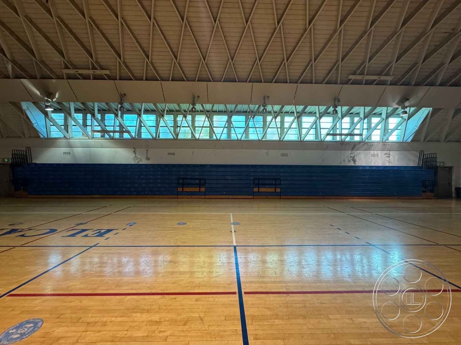 Old School - suspended lighting fixtures, exposed wood ceiling beams, open floor plan layout, interior gym space, plain painted walls, hardwood gymnasium flooring, glazed windows for natural light, blue bleacher seating, vaulted ceiling structure, indoor sports court environment, red and blue court markings, accessible entries, sports facility design, broad recreational area, large window wall for ventilation, high-traffic athletic space