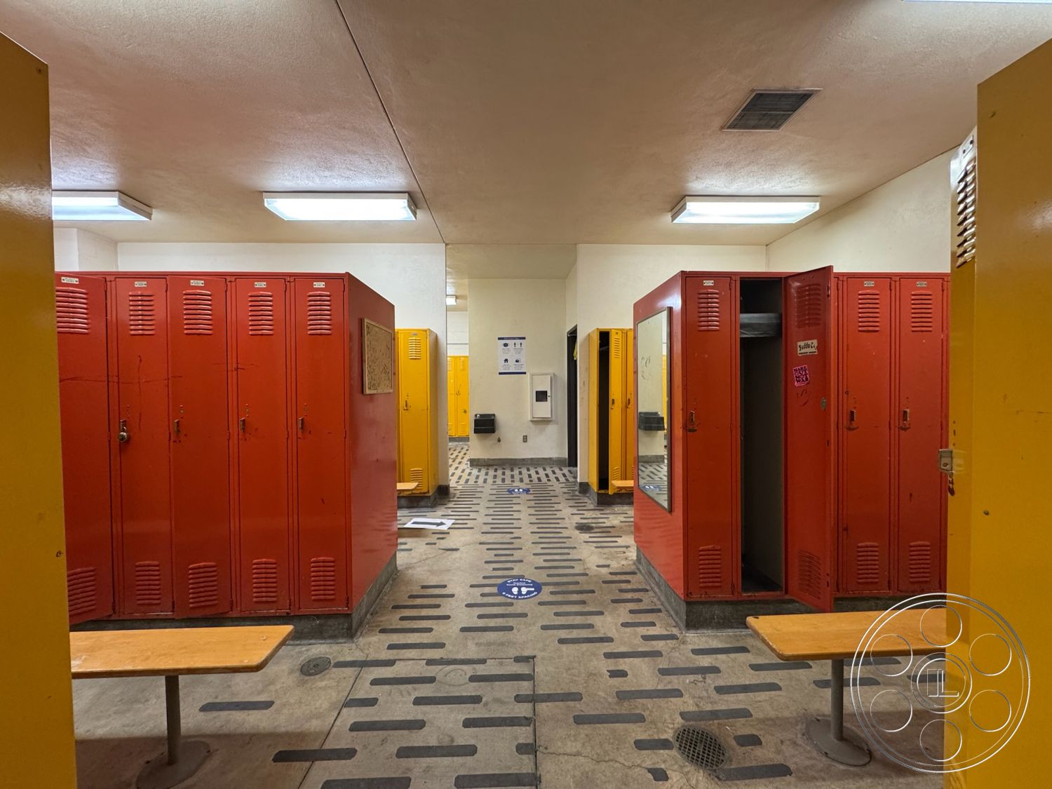 Old School - spacious layout, concrete floor, fluorescent lighting, textured tile flooring, athletic facility, changing area, decorative signage, interior locker room, open space configuration, red metal lockers, yellow locker doors, wood benches, walls with light paint, lockers with ventilation slats, mirrored wall section, school locker room