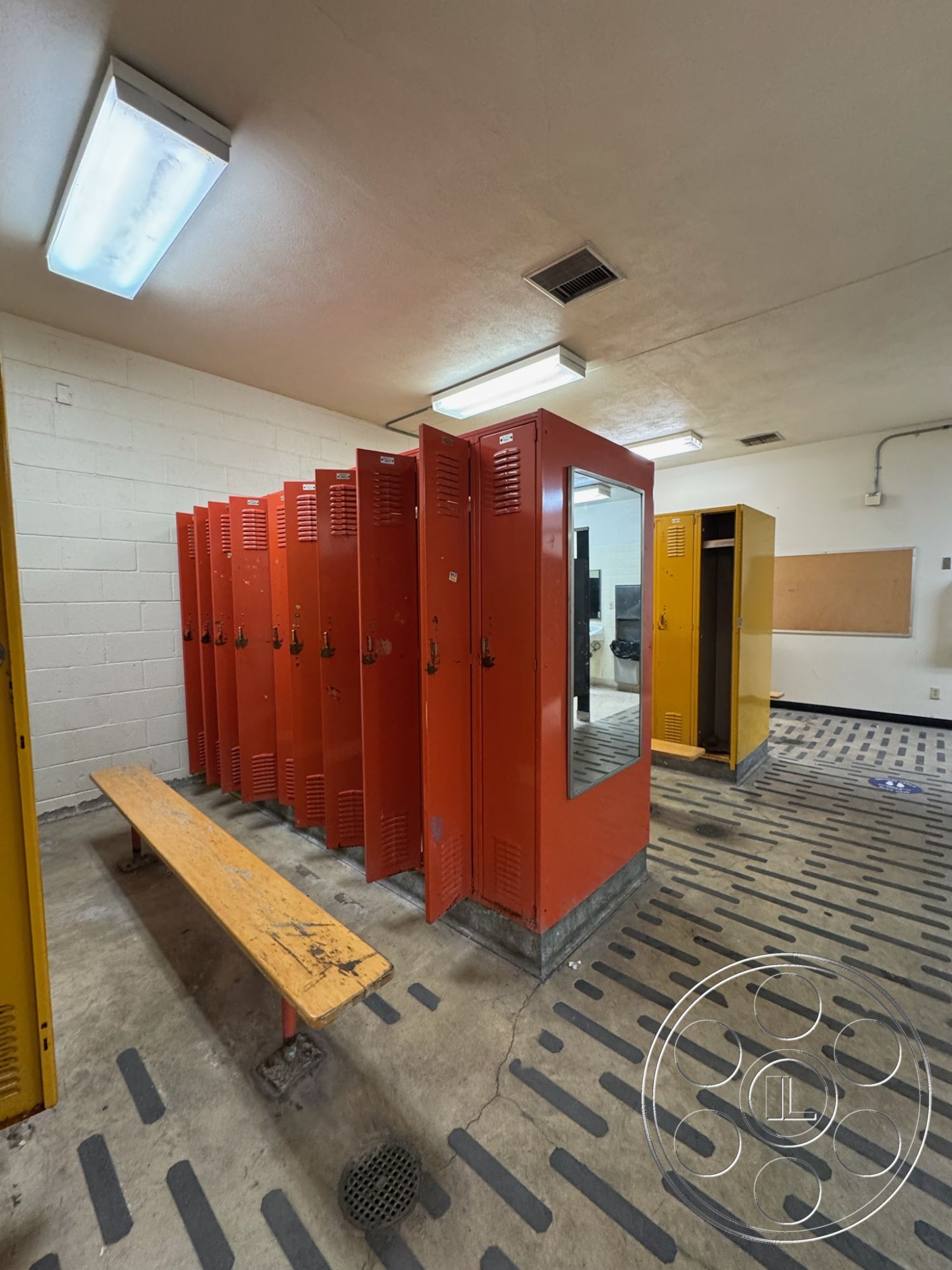 Old School - ventilation ducts, wood bench seating, fluorescent ceiling lights, interior locker room, metal lockers with mirrors, orange and yellow painted lockers, concrete floor with drainage grate, white painted cinder block walls, striped rubber flooring, lockers arranged in rows