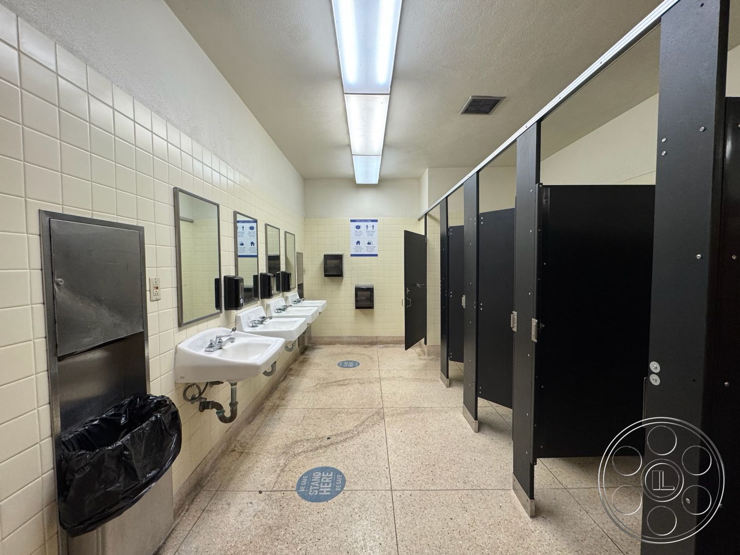 Old School - fluorescent ceiling lights, interior restroom design, glossy tile bathroom walls, porcelain sink countertops, stainless steel sink fixtures, laminate restroom partitions, black steel bathroom stalls, stainless steel trash receptacle, water-resistant linoleum flooring, mirrored wall above sinks, large wall-mounted soap dispensers, ADA-compliant restroom layout, modern commercial restroom, clean and minimalistic design, public restroom accessibility features