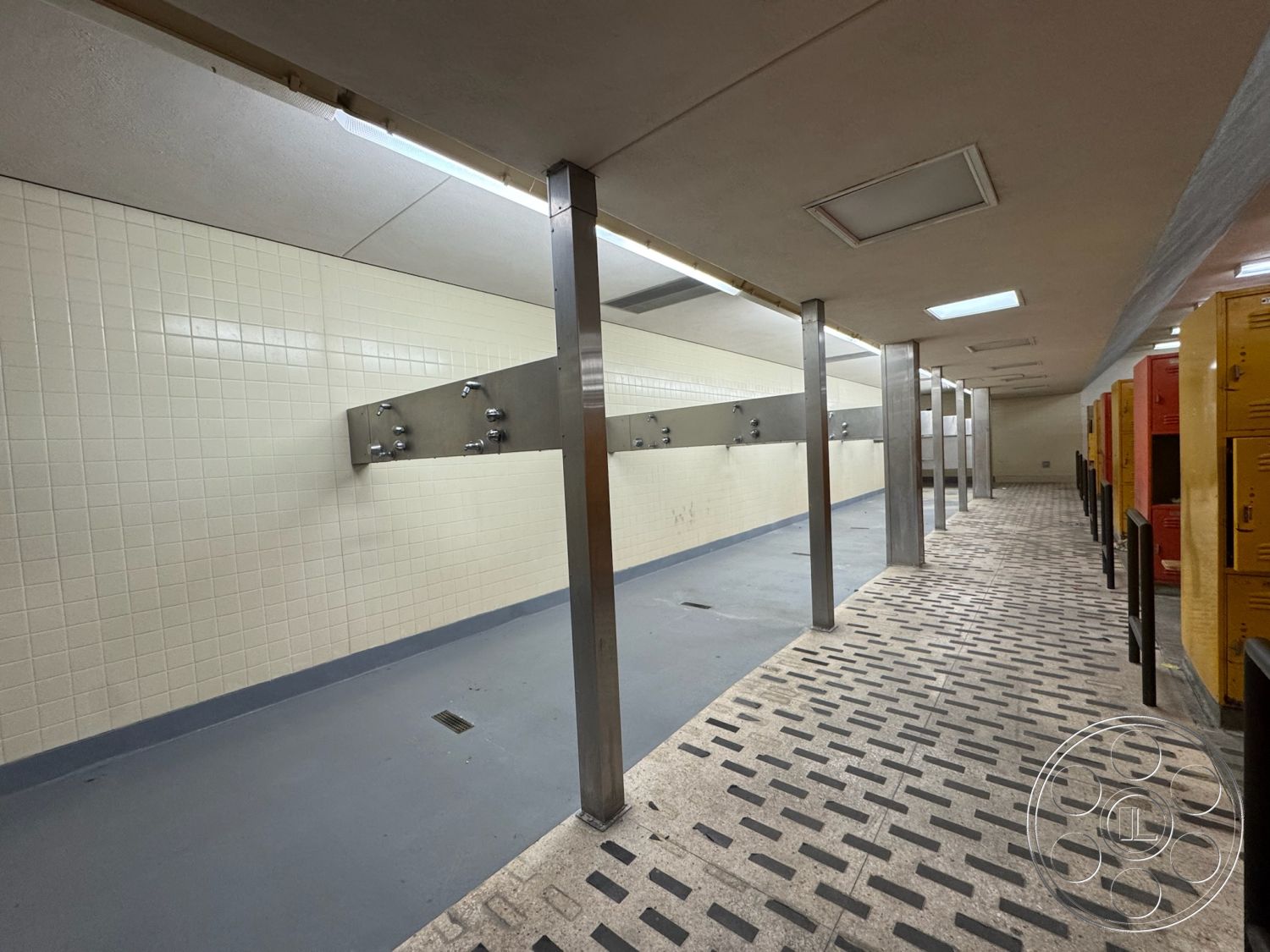 Old School - industrial design elements, metal support columns, exposed ceiling lights, interior shower area, non-slip flooring, white ceramic tile walls, steel shower fixtures, concrete floor with drainage, lockers with yellow and orange finishes, textured floor tiles, locker room layout, accessible shower area
