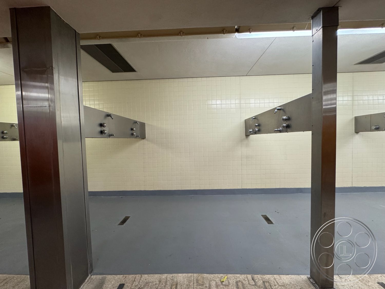 Old School - neutral color palette, minimalist design, gray vinyl flooring, stainless steel shower fixtures, flush-mounted ceiling lights, open shower layout, white ceramic tile walls, industrial shower area, commercial restroom interior, metal structural columns, water drainage floor grates, tiled shower rinse stations, high-traffic utility space