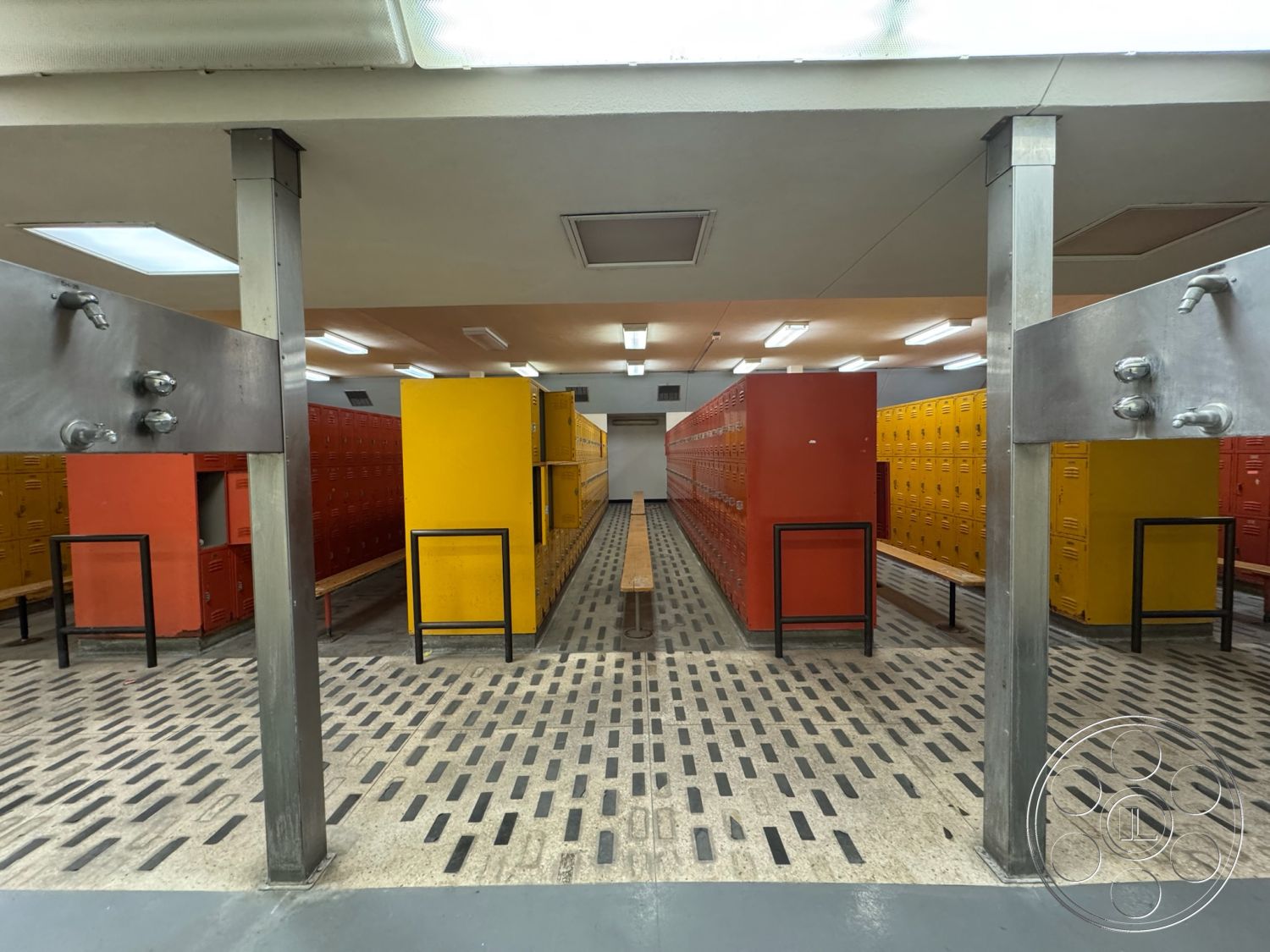 Old School - open layout, concrete flooring, functional design, industrial style, metal lockers, fluorescent ceiling lights, interior locker room, metal bench seating, yellow metal lockers, red metal lockers, textured concrete tile floor, gray steel support columns, shower area with wall-mounted faucets, amenity space, youth sports locker room
