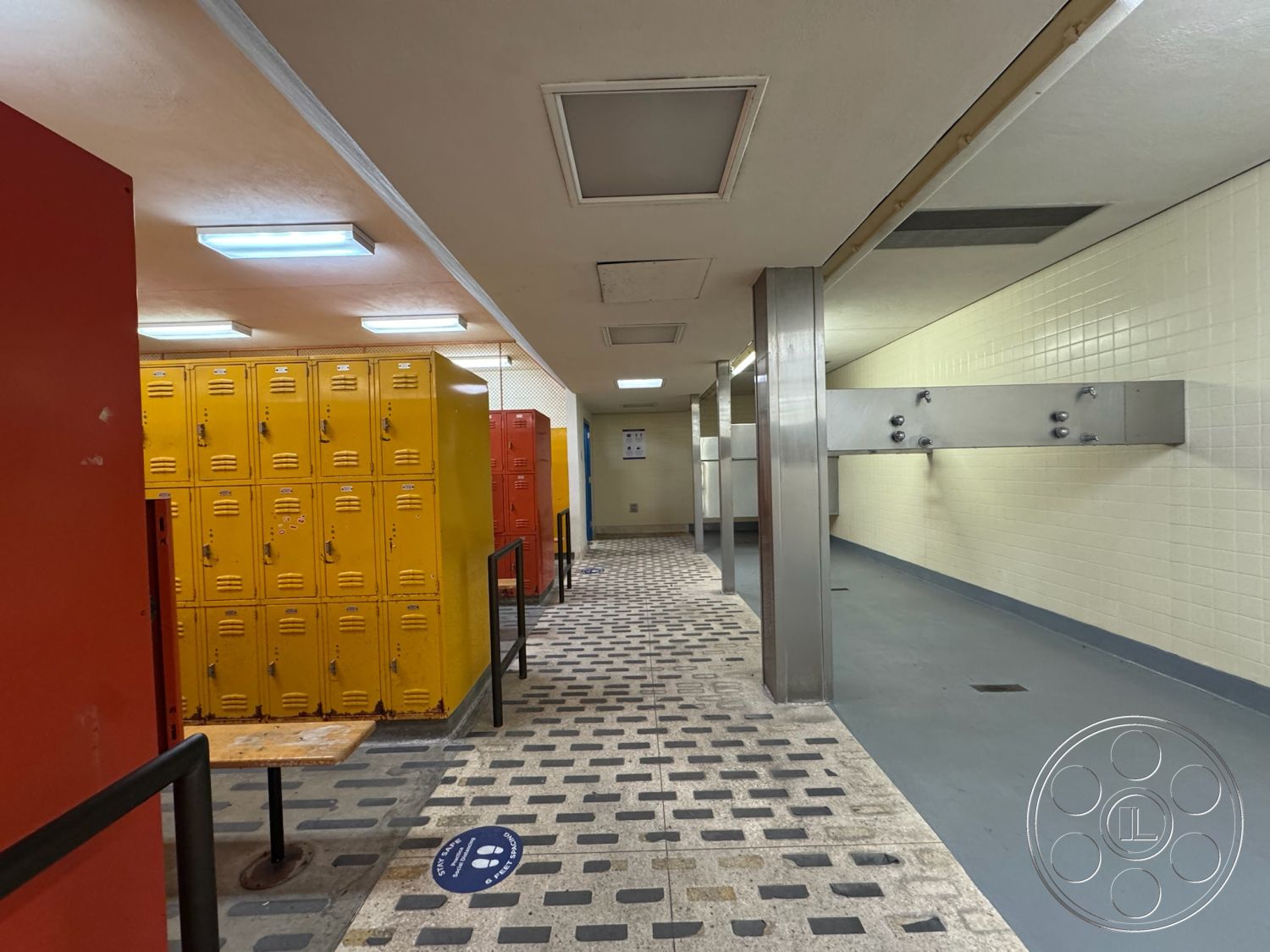 Old School - fluorescent lighting, exposed ceiling, white tiled walls, concrete floor finish, stainless steel shower fixtures, interior locker room, yellow metal lockers, red metal lockers, grey tiled flooring, patterned floor tiles, safety signage on floor, wooden table with metal legs