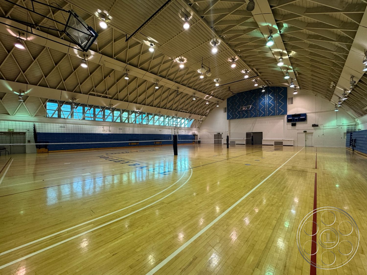 Old School - bleacher seating, hardwood gymnasium flooring, blue acoustic wall panels, sports facility interior, vaulted ceiling with exposed trusses, high ceiling with integrated lighting, glass windows with natural light, volleyball court markings, gym equipment storage, gymnasium space for multipurpose activities
