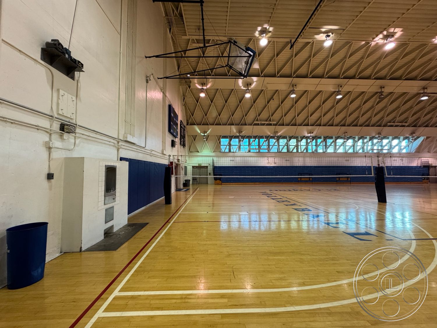 Old School - high vaulted ceiling, exposed metal beams, painted drywall walls, industrial style design, glass block windows, brightly lit environment, bench seating area, basketball court markings, wood gymnasium flooring, blue vinyl wall padding, indoor sports facility, large open space interior, concrete block storage area, urban sports center setting