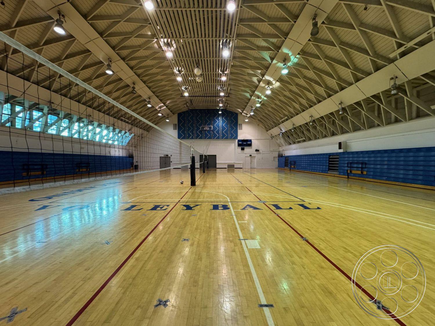 Old School - open layout design, glass block windows, hardwood gymnasium flooring, bright overhead lighting fixtures, volleyball court markings, blue vinyl wall padding, exposed wooden truss ceiling, indoor sports arena setting, multi-purpose athletic space, high-ceiling structure, painted gymnasium walls, bleacher seating arrangements, interior sports court environment