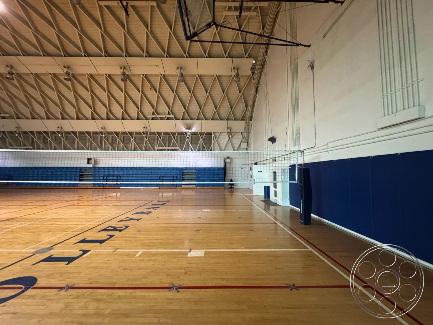 Old School - recreational space, interior sports facility, blue vinyl wall padding, hardwood gymnasium floor, high vaulted ceiling with exposed wooden trusses, painted white interior walls, volleyball net with metal posts, gym bleachers along one wall, dimensional lighting fixtures on ceiling, marked volleyball court lines on floor, acoustic wall panels for sound dampening, community gymnasium setting