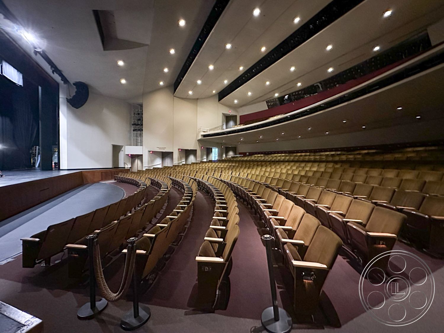 JT Theatre - modern exit signage, exposed structural beams, wooden theater seats, theater interior, sound system integration, backlit signage, open performance area, curved auditorium seating, black acoustical ceiling panels, spotlights and recessed lighting, stage area with black curtain, carpeted auditorium floor, multiple seating rows, contemporary theater design, accessible seating sections, wall-mounted speaker system, foyer with box office, audience viewing angles, emergency exit pathways