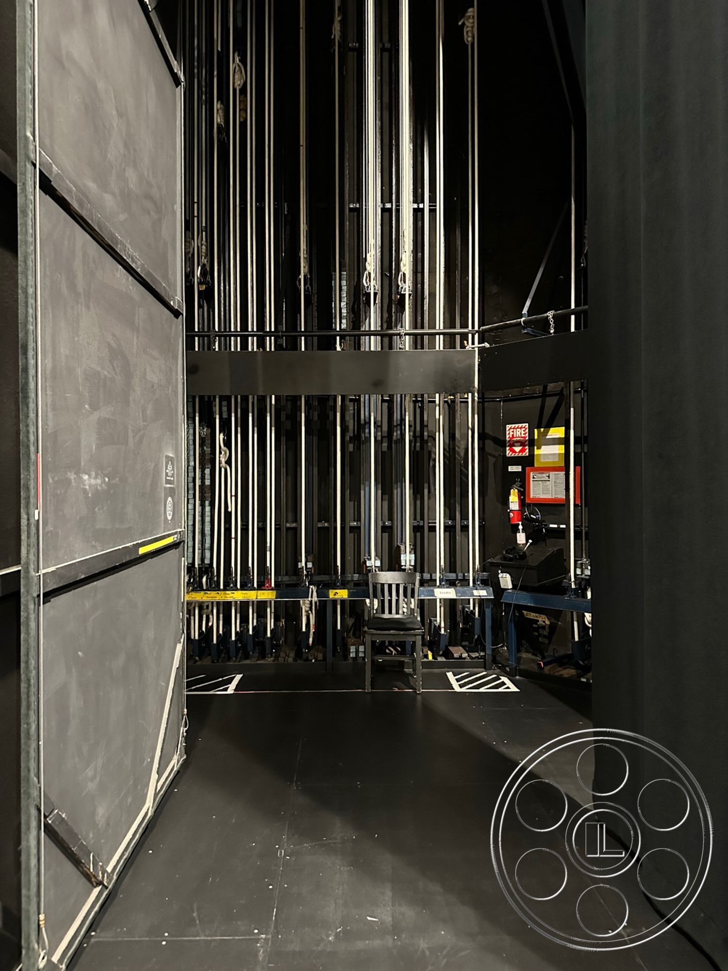 JT Theatre - backstage access, black painted walls, black stage floor, fire safety signage, wooden chair on stage, interior stage area, metal rigging system, plywood stage elements, exposed lighting rig, stage left wing, manual fly system, show control panel, theater production environment, adjacent control booth, audience seating area access