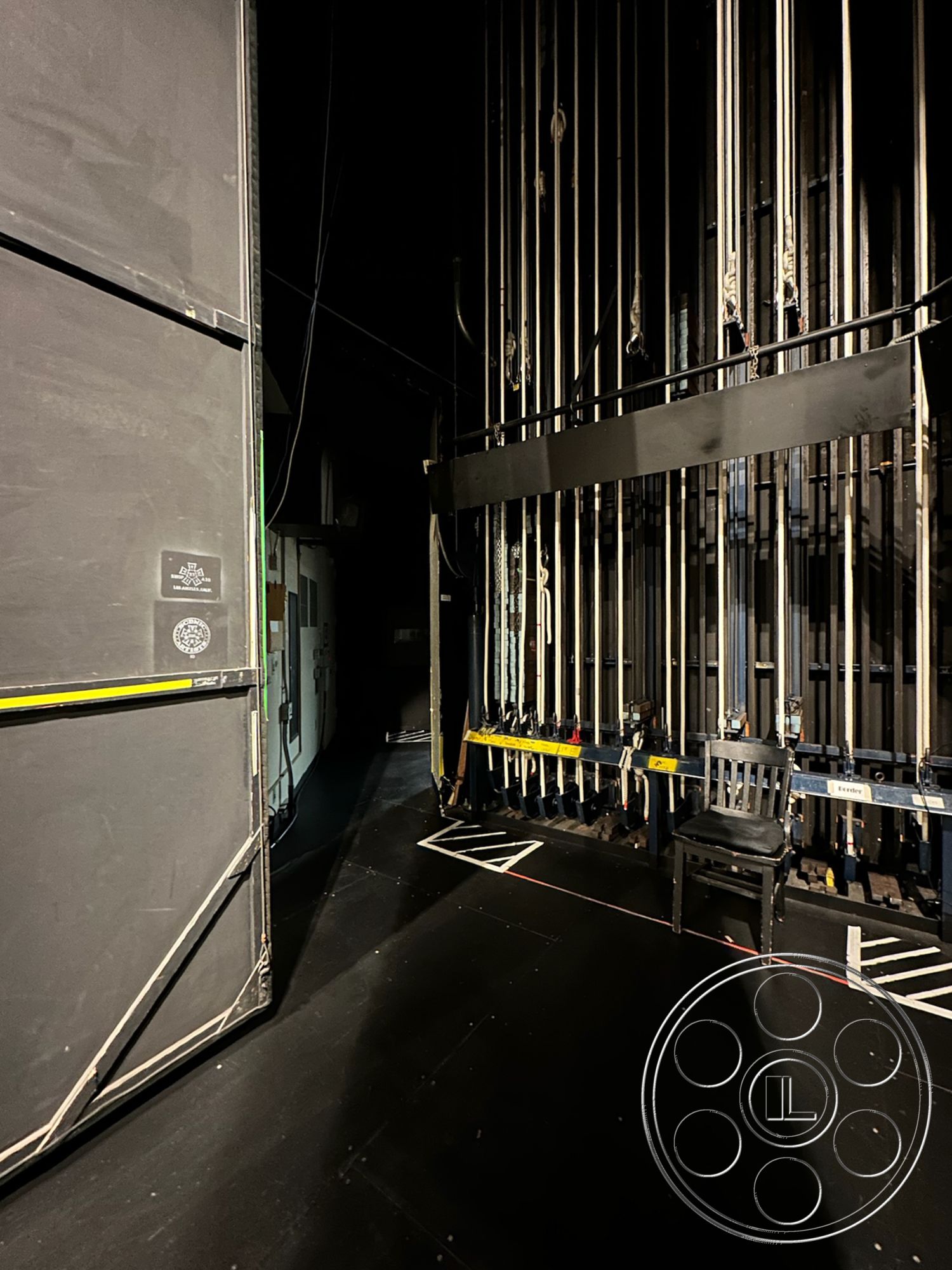 JT Theatre - dark color scheme, exposed lighting fixtures, backstage access corridor, acoustic paneling on walls, emergency exit signage, black painted stage walls, hardwood theater flooring, industrial metal rigging system, studio backdrop curtains, theatrical prop storage area, black box theater interior, stage management console area, director's viewing area, flexible performance space, seating area for crew, caution tape floor markings, movable scenery tracks, ambient stage lighting, technical equipment space, high ceiling with rigging