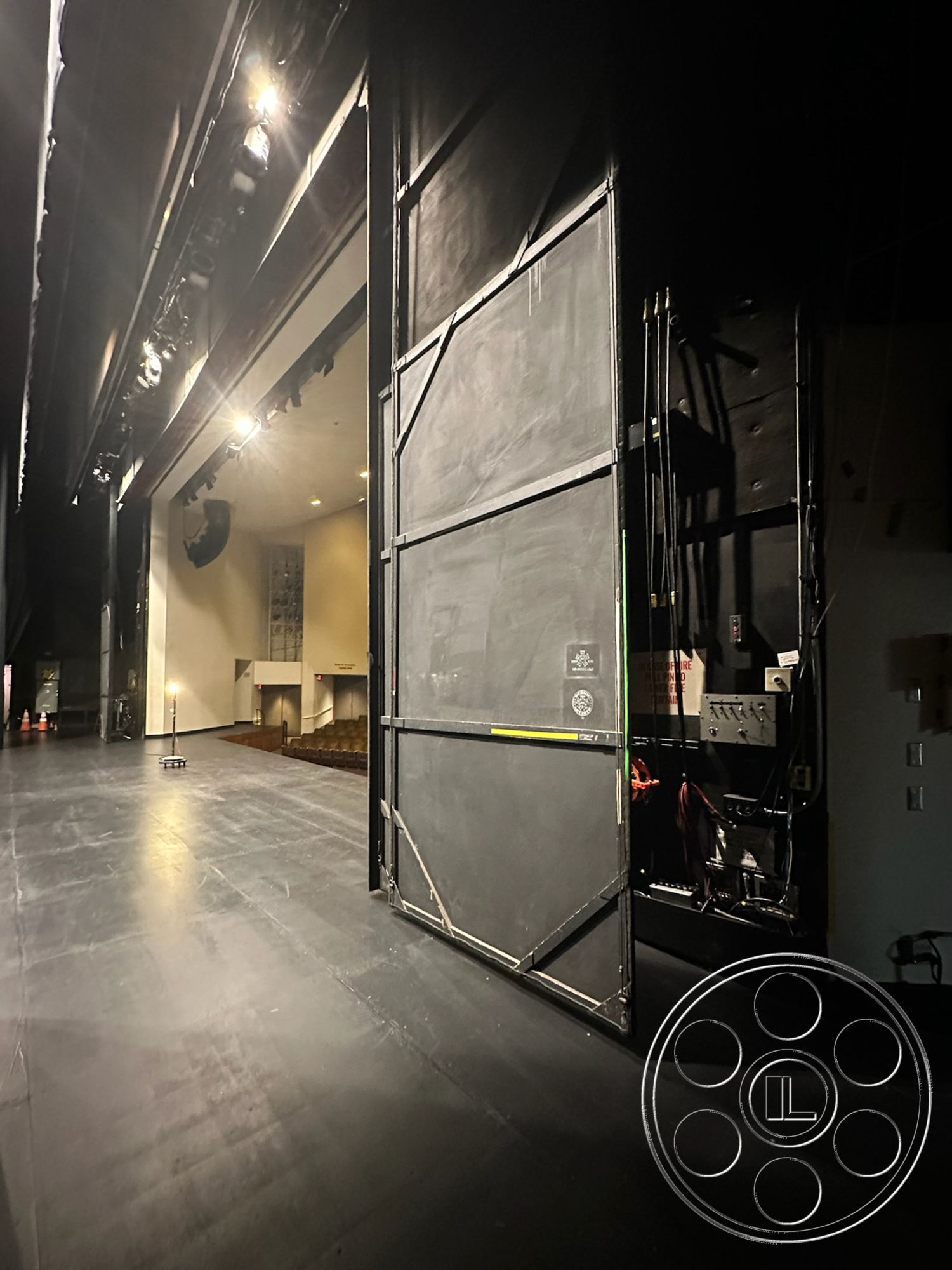 JT Theatre - auditorium seating in background, interior stage flooring, black stage floor panels, theater backdrop, dimly lit theater interior, professional stage lighting fixtures, curtained stage entry, sound equipment control panel, exposed stage curtain track, backstage work area, industrial theater structural elements, smooth polished floor surface, dark-painted wall surfaces