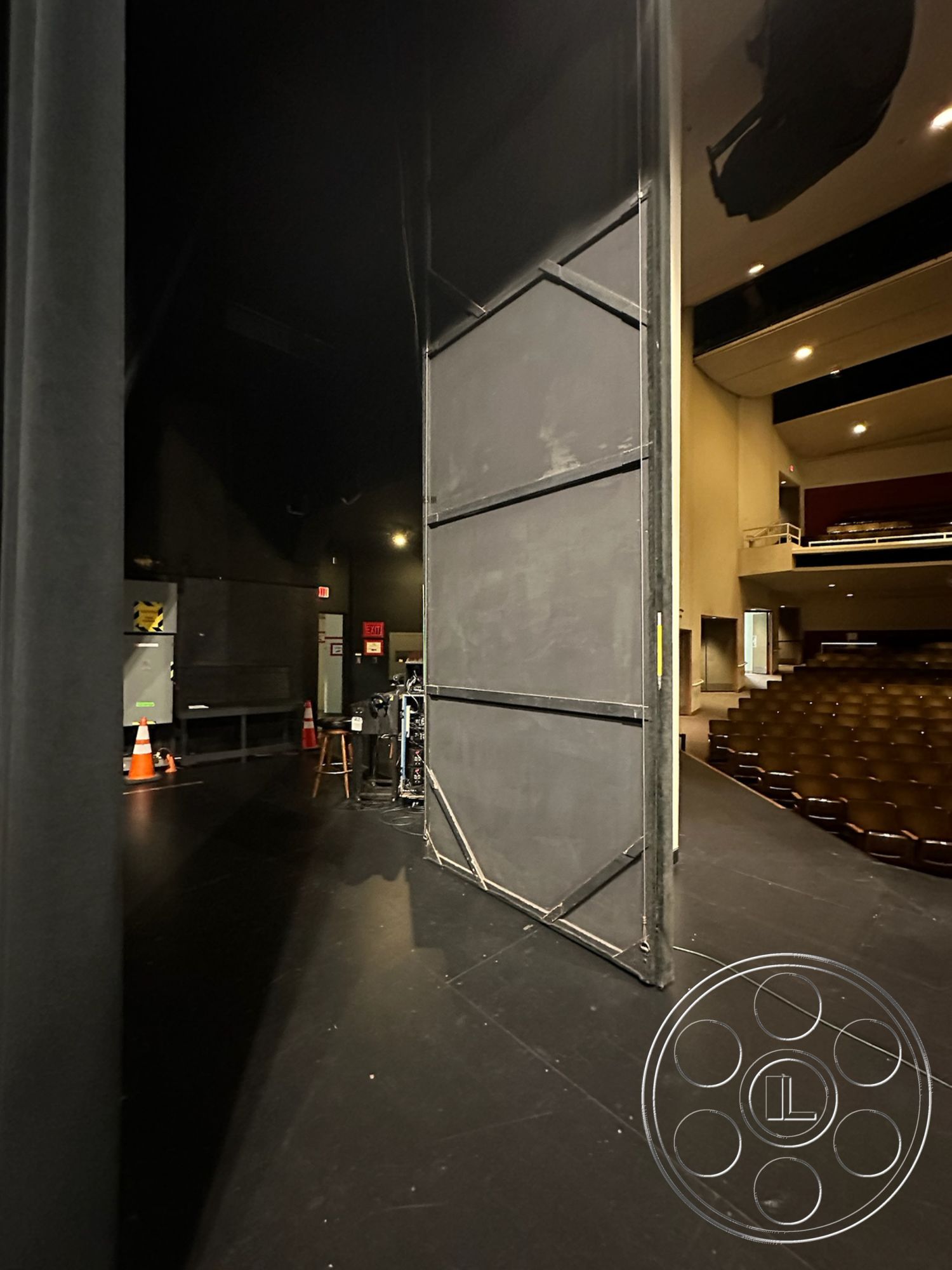 JT Theatre - raised seating area, black painted walls, acoustic panels, black stage curtain, wooden stage floor, theater auditorium seating, metal stage structure, spotlights on stage, backstage environment, exit signs, stage left, performance venue interior, control booth access, auditorium entrance, theater lighting setup