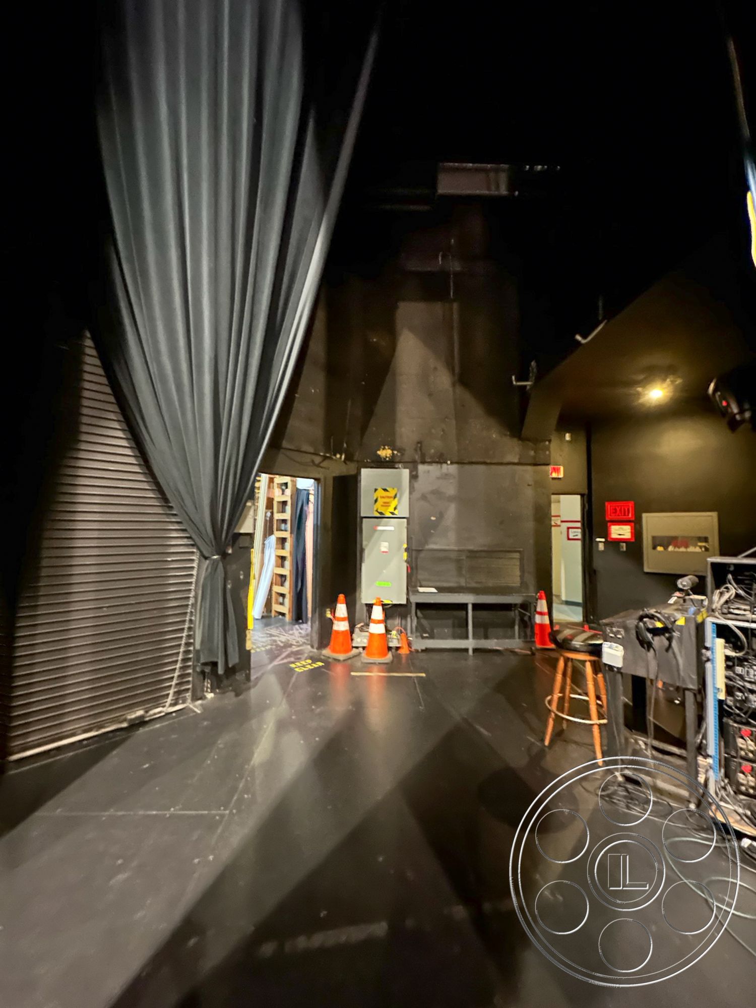 JT Theatre - multi-purpose space, folding chair, concrete block wall, dimly lit interior, black stage flooring, exposed metal structural elements, metal ladder, utility storage area, theater production environment, performance venue interior, curtained stage entrance, industrial backstage area, lighting control station, sound equipment setup, staging area with chairs, safety cones in transit area, dark wood stool, electrical panel access, backstage exit signage, draped black curtains