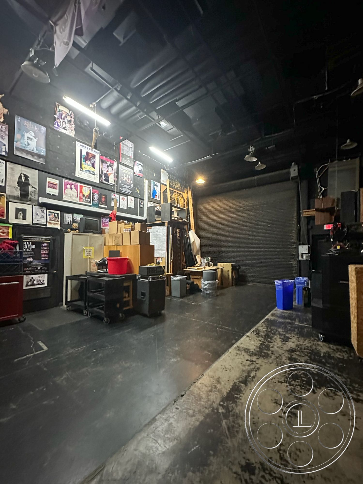 JT Theatre - fluorescent ceiling lights, urban industrial setting, metal storage cabinets, plastic storage bins, exposed brick accent walls, black-painted ceiling tiles, concrete industrial flooring, artistic poster wall decor, open workspace interior, rolling carts for equipment, loading dock garage door, utility area with discarded materials, functionally designed layout, multi-purpose studio space