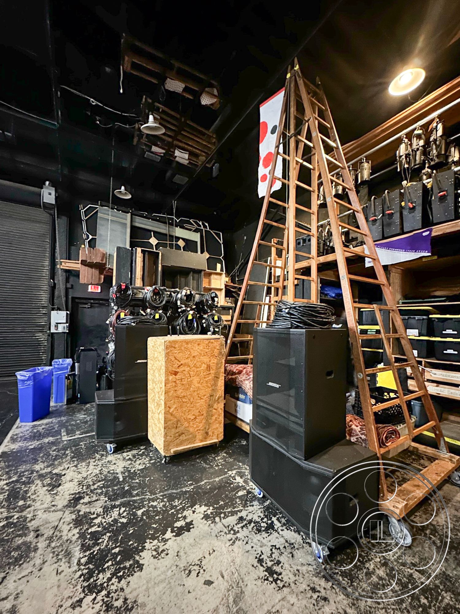 JT Theatre - exposed ceiling beams, stacked speaker cabinets, wood shelving unit, interior industrial space, black painted concrete walls, wooden ladder structure, steel audio equipment, rollers on sound system boxes, plywood storage box, lighting fixtures overhead, plastic garbage bins, hardwood floor panels, electrical equipment storage, active workspace, temporary event storage area