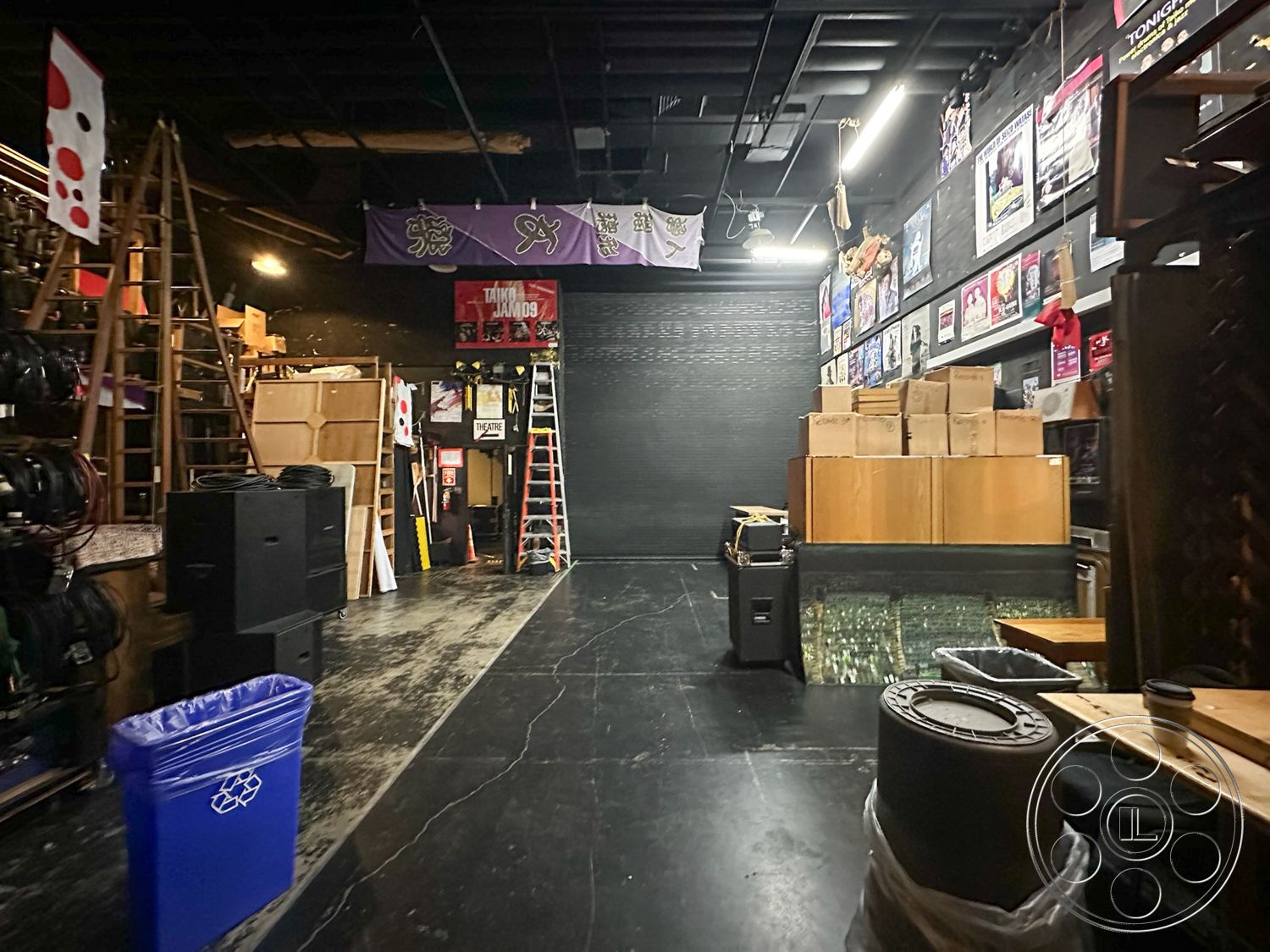 JT Theatre - concrete floor, creative workspace, open space layout, metal shelving units, industrial lighting fixtures, recycling bin, stacked cardboard boxes, sound equipment storage, exposed black painted walls, wooden storage ladders, black acoustic panels, theatrical posters on walls, coffee cup on table, community performance venue, urban arts setting