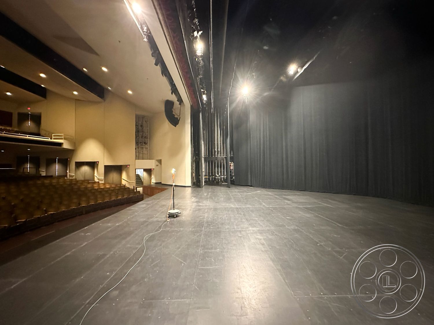 JT Theatre - exposed lighting fixtures, dark wood paneling on walls, acoustic panel walls, black stage flooring, curved theater seating, velvet stage curtains, backstage area with wooden support beams, auditorium ceiling with recessed lighting, sprung performance stage floor, spotlight lighting overhead, entrance lobby with glass doors, theater box with plush seating, interior auditorium design, theater sound equipment setup, modern performance art space, seating rows with fabric upholstery