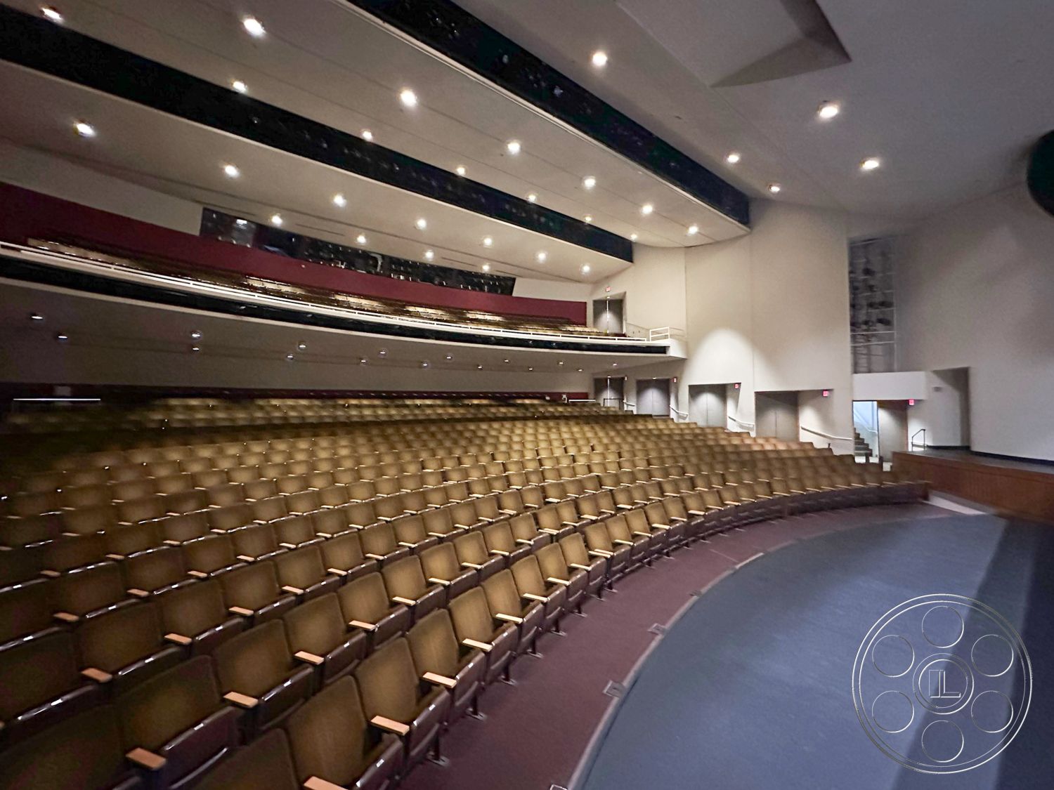 JT Theatre - warm wood accents, recessed ceiling lighting, black stage curtains, auditorium interior, acoustic wall treatment, curved seating layout, theater seating arrangement, upholstered theater seats, dark wood stage platform, stage lighting fixtures, single-level auditorium, floating stage edge, gray acoustic wall panels, high ceiling with sound dampening, built-in railing for audience safety, wide entry doors, accessible seating options, elevated seating tier, colorful balcony trims, sound equipment area, emergency lighting system