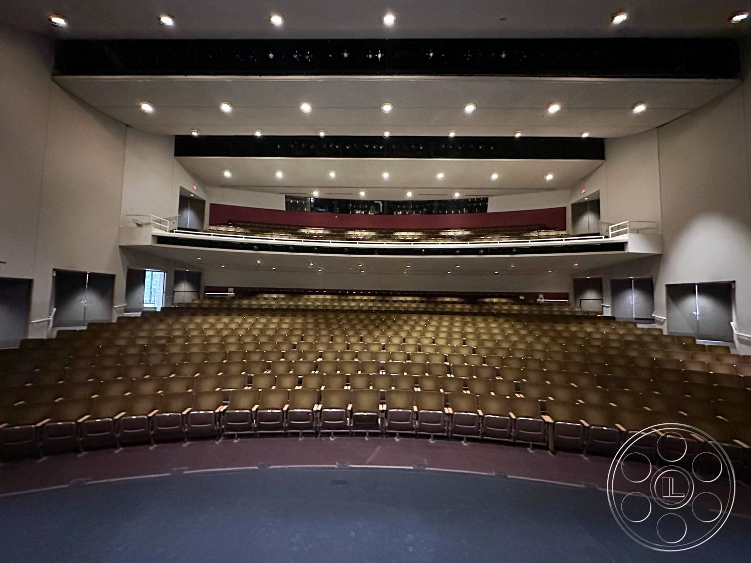 JT Theatre - flush ceiling lights, neutral painted walls, high ceiling design, interior theater seating, acoustic panel walls, curved auditorium seating arrangement, multi-level seating tiers, emergency exit signage, spotlight lighting fixtures, accessible seating options, raised stage area, black stage curtain backdrop, modern auditorium design, orchestration pit, comfortable padded chairs, horizontal balcony viewing area, steel handrail balcony, sound system speaker setup, entrance doors to auditorium, lobby area adjacent to theater