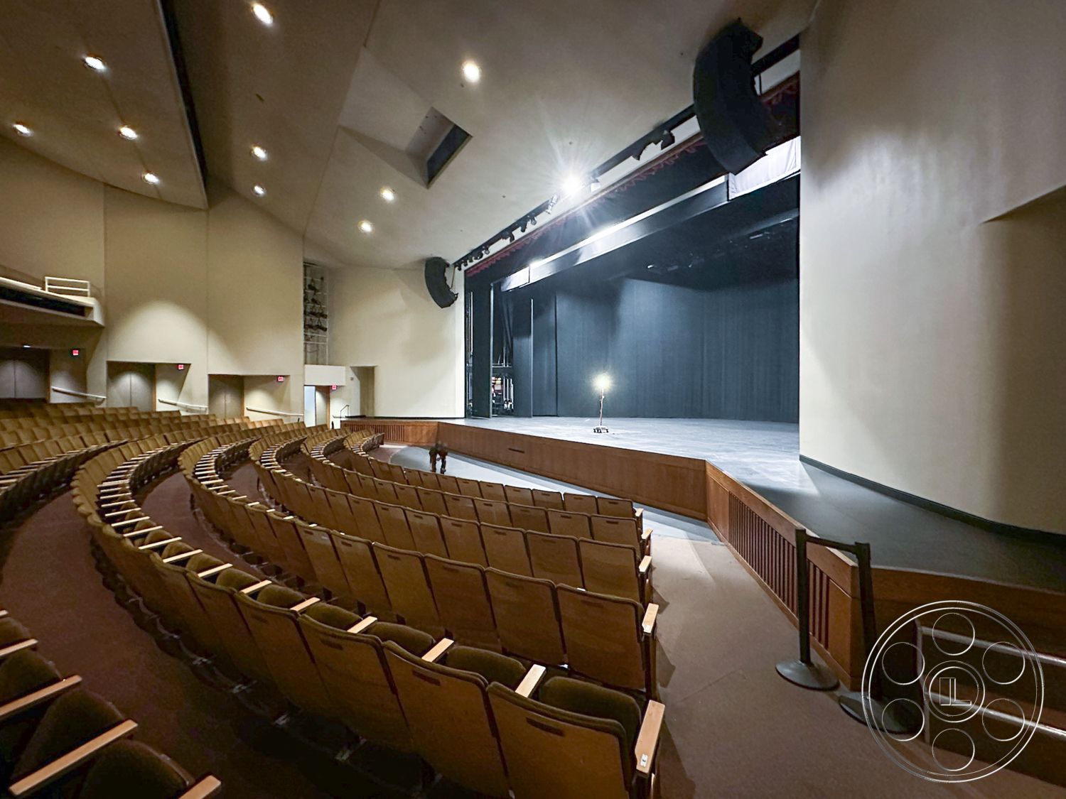 JT Theatre - acoustic ceiling panels, textured plaster wall, curved seating arrangement, wood paneling accents, wooden auditorium seating, stage with black curtains, circular theater layout, high-vaulted ceiling, theatrical lighting fixtures, darkened performance stage, exposed beam architectural details, composite stage flooring, soundproof wall treatment, auditorium lobby entrance, interior theater space, raised platform stage, auditorium exit signs, emergency exit stairs, theater sound systems, architectural design elements