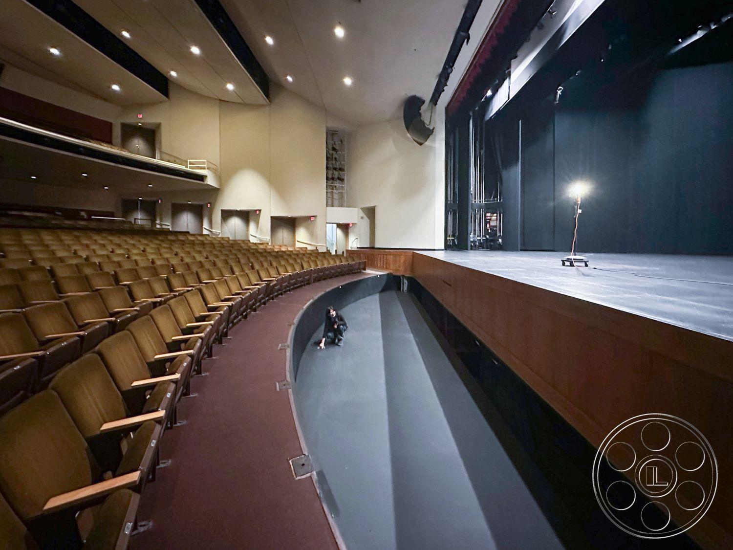 JT Theatre - recessed lighting fixtures, acoustic ceiling panels, neutral wall treatments, backstage access doors, sound system speakers, auditorium interior, black stage curtain, multi-tiered seating arrangement, wooden seating rows, continuous stage platform, curved theater floor, dark wood stage border, red fabric acoustic panels, spotlight lighting system, theater performance space