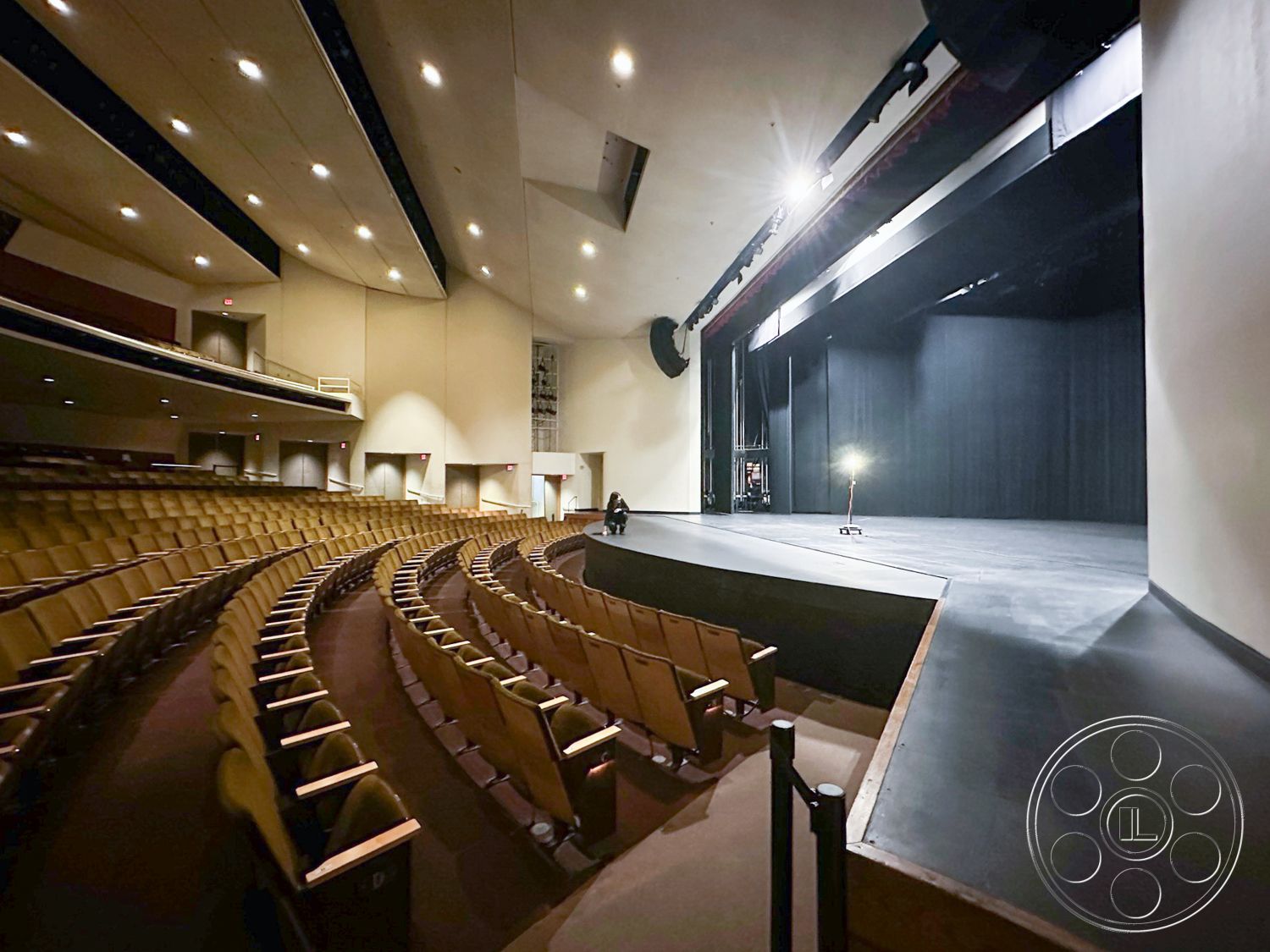 JT Theatre - recessed ceiling lighting, exposed structural beams, acoustic wall panels, backstage access doors, interior auditorium space, emergency exit signage, wooden stage floor, tiered seating with upholstered chairs, curved row seating arrangement, black stage backdrop, control booth at rear with glass window, high ceiling with integrated sound system, dimmable house lighting, theater-style sound isolation, multi-use performance space, performance stage with a slight incline, municipal theater design, modern auditorium aesthetics, public performance venue, dynamic lighting capabilities