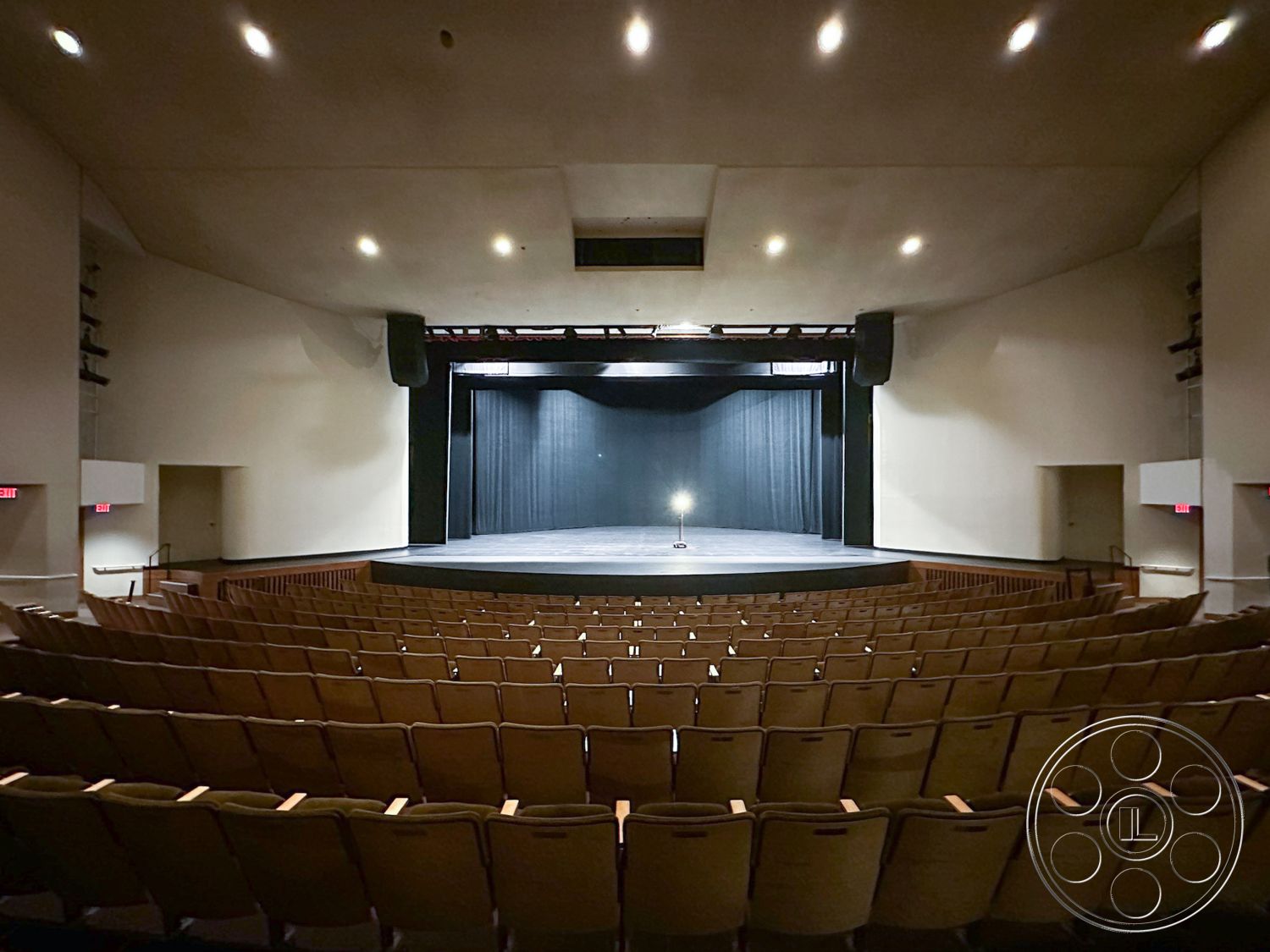 JT Theatre - high ceiling with recessed lighting, acoustic panel walls, backstage access doors, sound system speakers, wooden auditorium seating, entrance lobby area, black stage curtain, stage lighting fixtures, raised platform stage, interior theater auditorium, curved stage, drum-shaped ceiling, staircase to the stage, multimedia projection setup, performance space ambiance