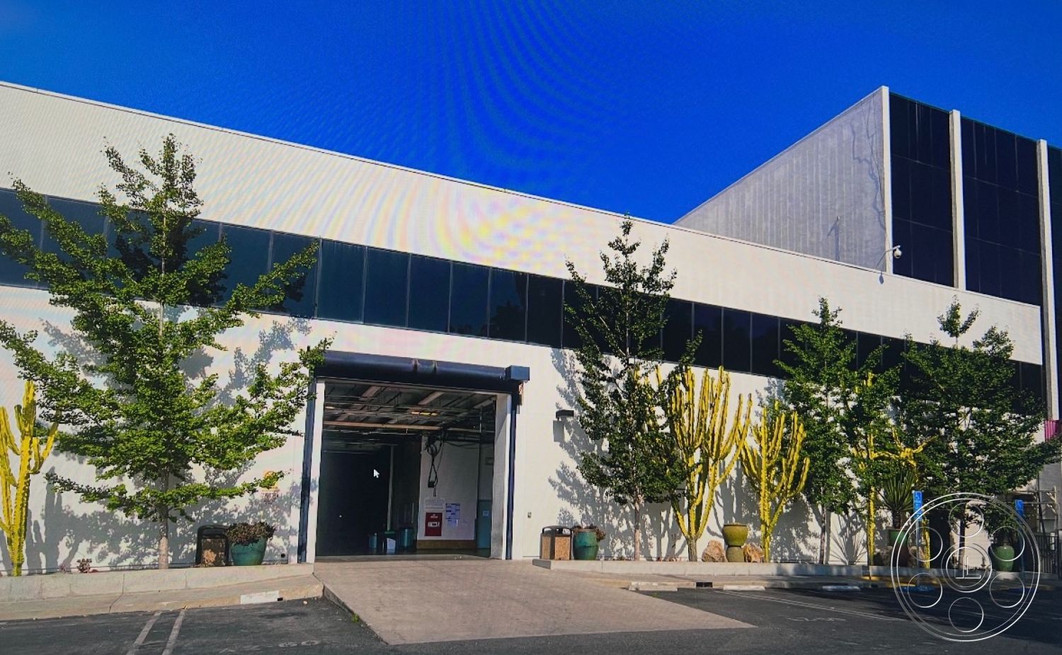 Stage 33 - exterior, modern architectural style, steel frame structure, industrial building, large loading dock, flat roof design, concrete façade, glass storefront windows, paved parking area, urban commercial environment, landscape with desert plants, basketball court tile surface, potted cacti and trees, open garage entrance, blue sky backdrop