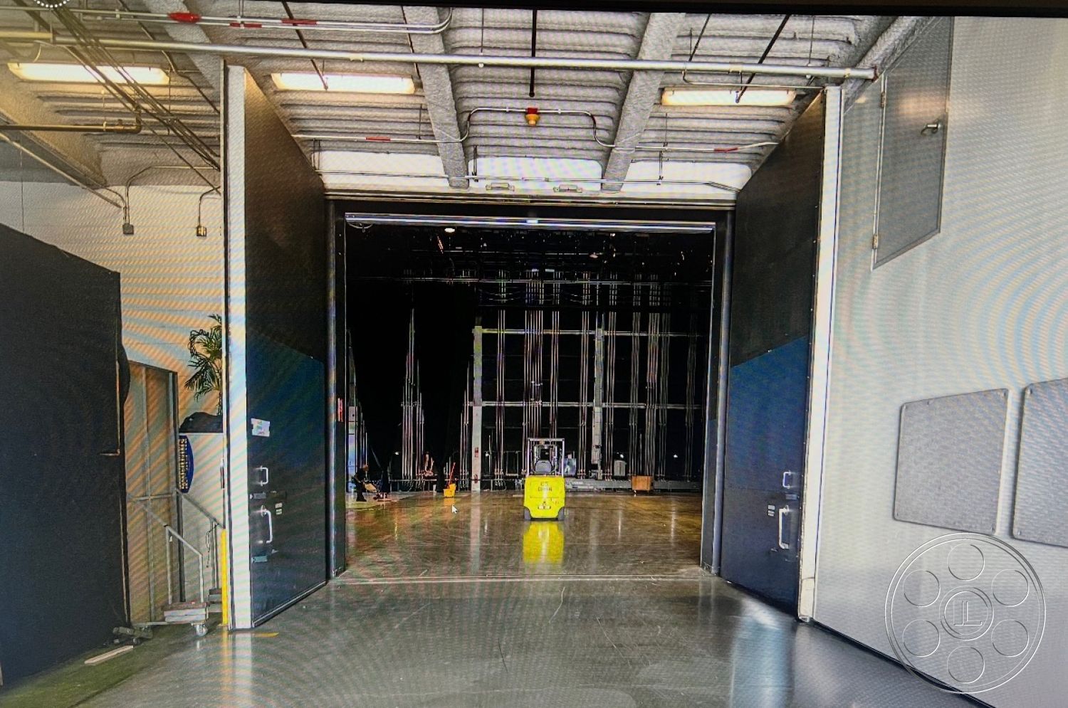 Stage 33 - polished concrete floor, modern design elements, functional entryway, industrial space, acoustic wall treatment, high ceiling with exposed ductwork, large loading doors, metal frame doors, large opening to performance area, dim lighting with fluorescent fixtures, open rehearsal space, curved wall panels, flexible space for events, unfinished warehouse aesthetic, adjacent storage area, functional space for production