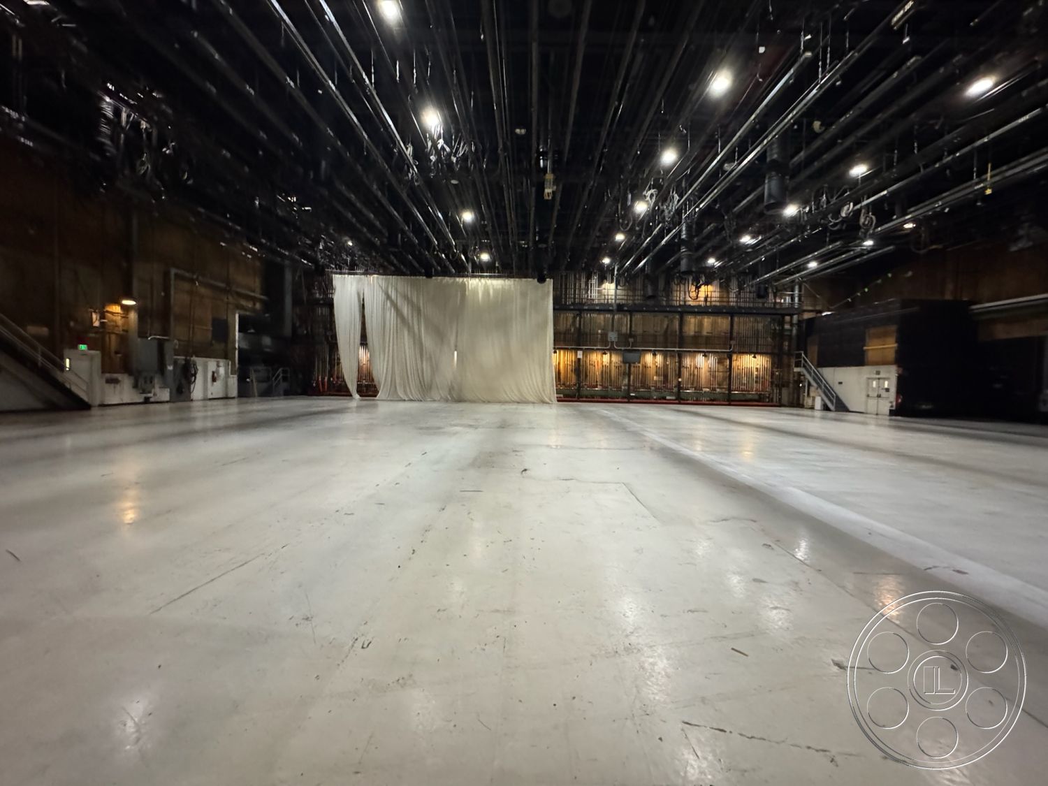 Stage 33 - rehearsal space, overhead lighting grid, sound stage, large open area, exposed industrial ceiling, interior studio space, high ceiling design, acoustic wall treatments, white painted concrete floor, curtained backdrop area, metal piping infrastructure, stage area for performances, dimly lit studio environment, flexible layout space, warehouse-style architecture, film production studio