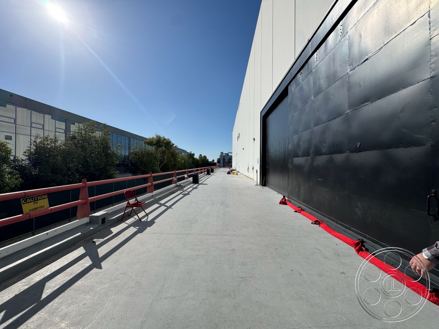Stage 33 - exterior, surrounding greenery, concrete walkway, minimalist architectural style, open sky view, sunlit urban setting, industrial building facade, black steel overhead door, metal railing with red accents, glass window sections, concrete surface finish, maintenance access area, construction site caution signage, flat horizontal plane