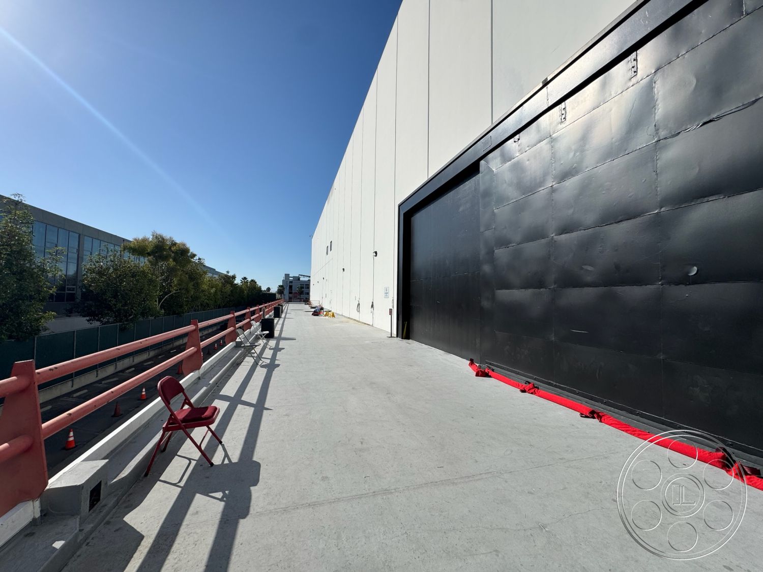 Stage 33 - outdoor seating area, concrete pavement, open outdoor space, flat roof design, exterior concrete walkway, straight pathway, minimalistic architectural style, large industrial black metal door, red metal railing, modern industrial building facade, landscaped area with trees, overcast urban setting, adjacent commercial buildings, hardscape adjacent to green space, warehouse building exterior