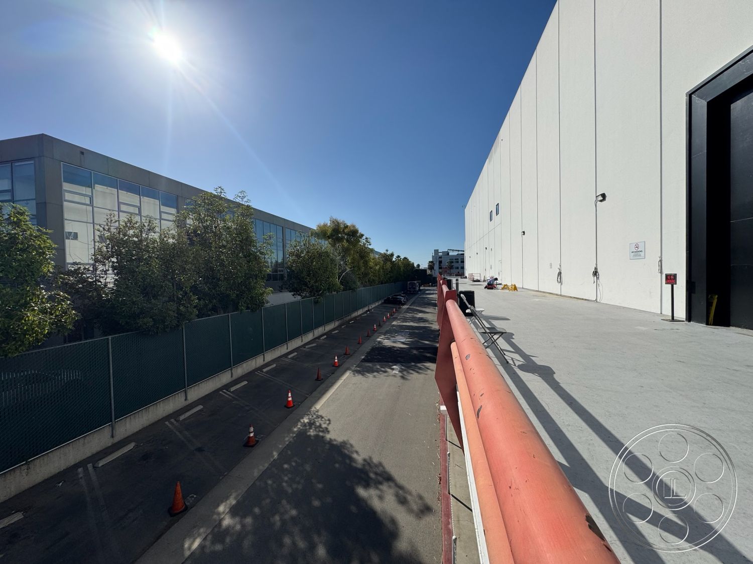 Stage 33 - surrounding urban environment, clear blue sky view, urban industrial setting, asphalt road surface, concrete parking area, single-story commercial building, industrial warehouse structure, exterior concrete loading dock, glass facade commercial building, painted white concrete walls, vegetated landscaping, chain link fence perimeter, sunlit open space, red-painted metal railings, outdoor logistical space