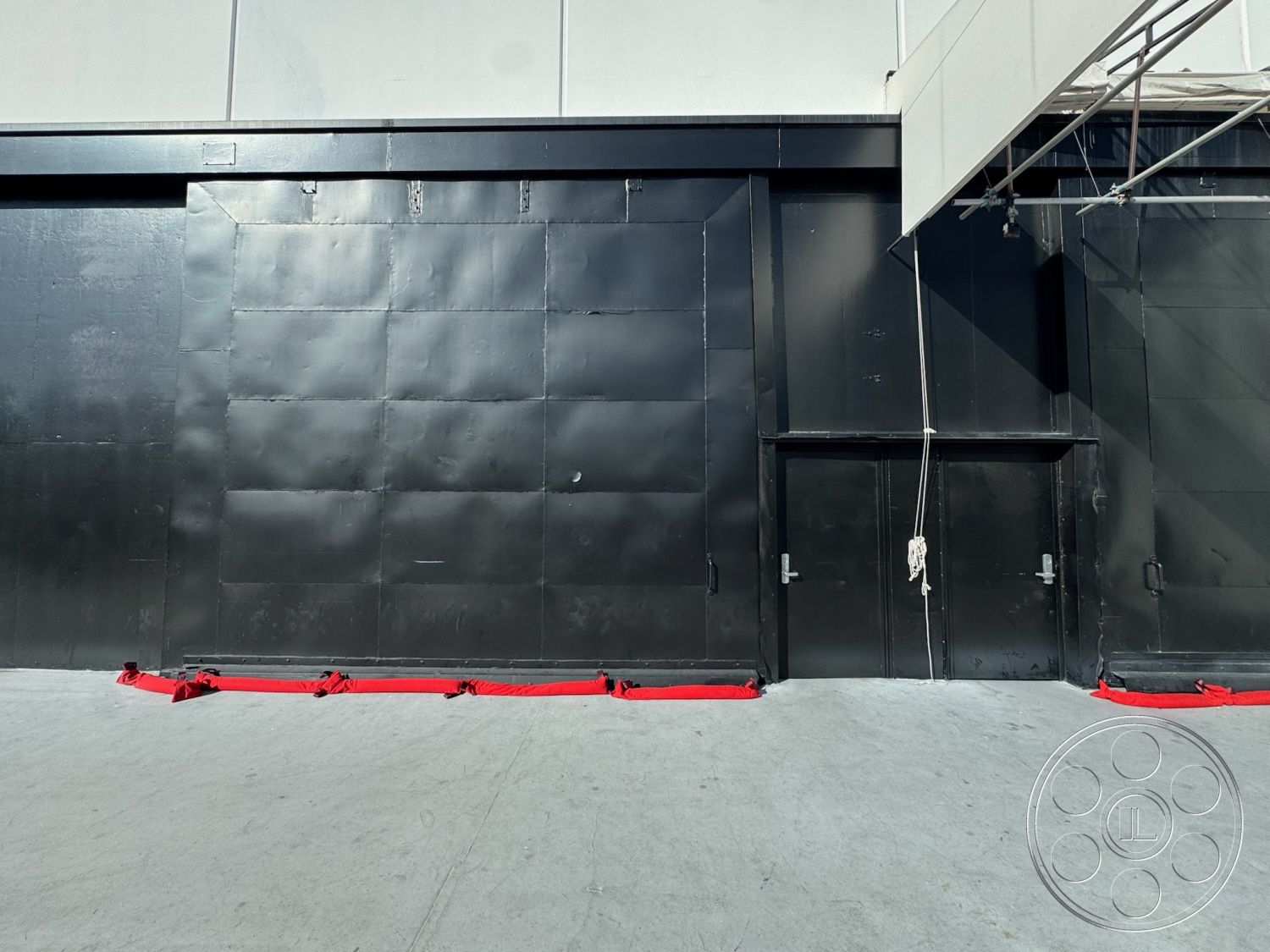 Stage 33 - concrete flooring, industrial design elements, exposed structural elements, minimalistic architectural style, black metal wall exterior, grid pattern wall panels, hidden door entrance, textured metal surface, red fabric barrier, large blank wall, scenic lighting setup potential
