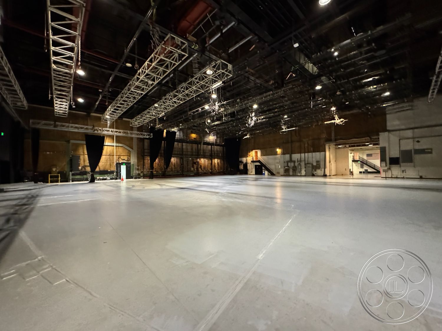 Stage 33 - open floor plan, black curtain stage backdrop, exposed concrete floors, warehouse-style interior, sound stage environment, industrial ceiling beams, high ceiling studio space, metal grid lighting system, commercial production studio, adjustable lighting grid, whitewashed concrete walls, exterior loading dock access